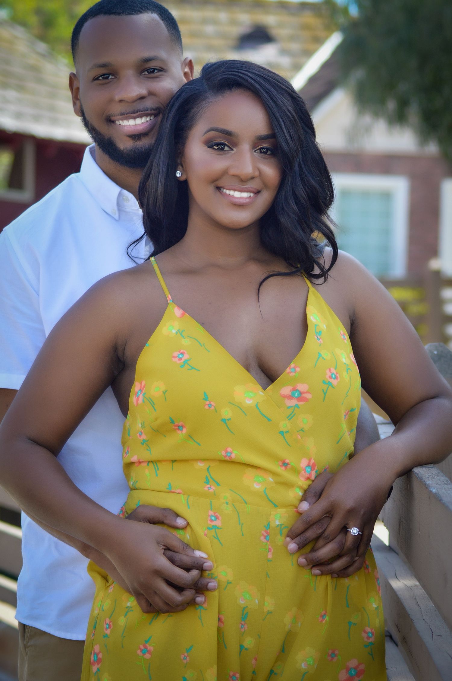 A smiling couple poses outdoors, woman in yellow floral dress, man in white shirt, both looking happy and embracing.