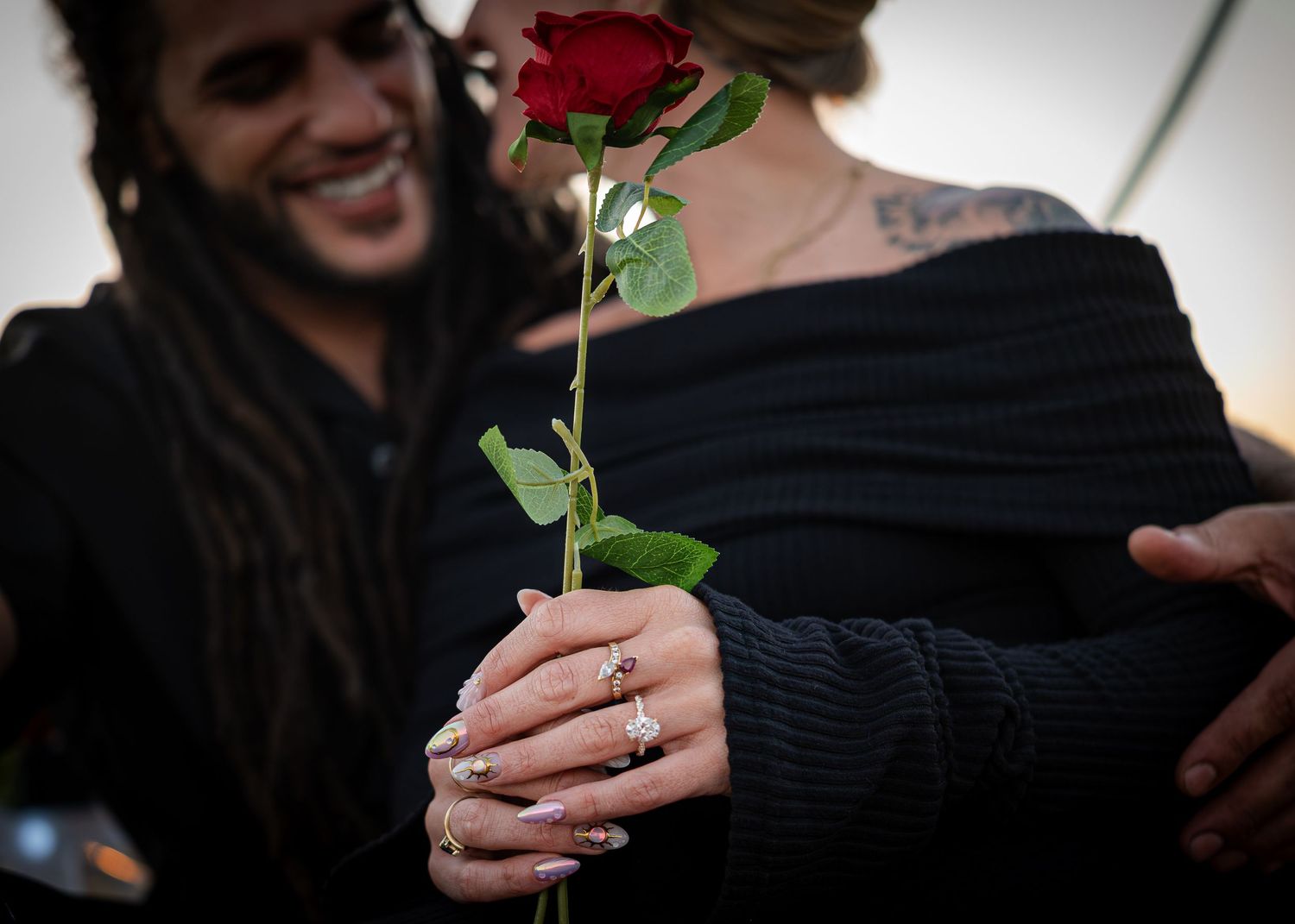 A couple in black clothing shares a romantic moment with a red rose, featuring tattooed skin and silver rings.
