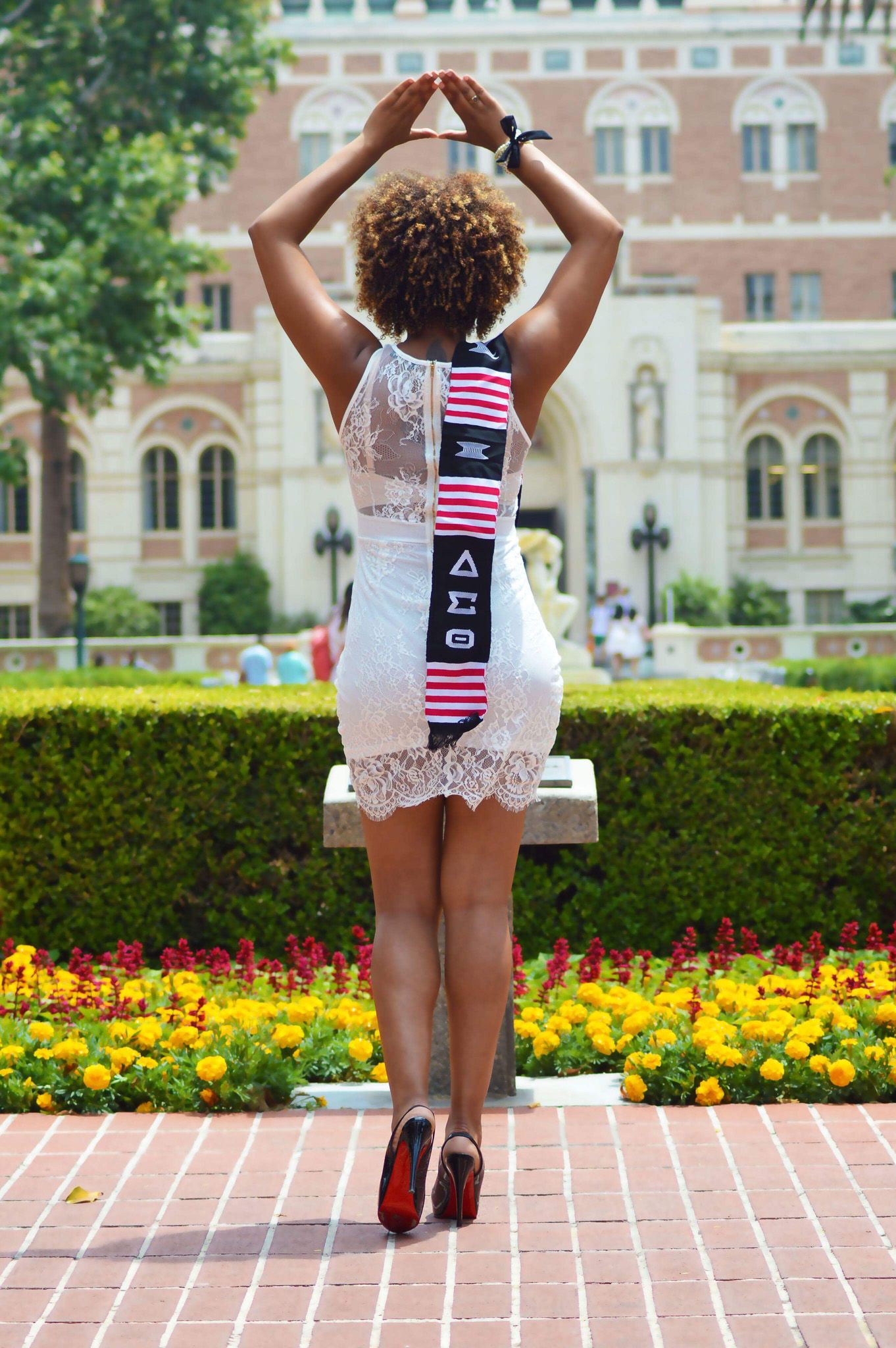 Graduate in white lace dress forms triangle pose showing graduation stole against campus building.