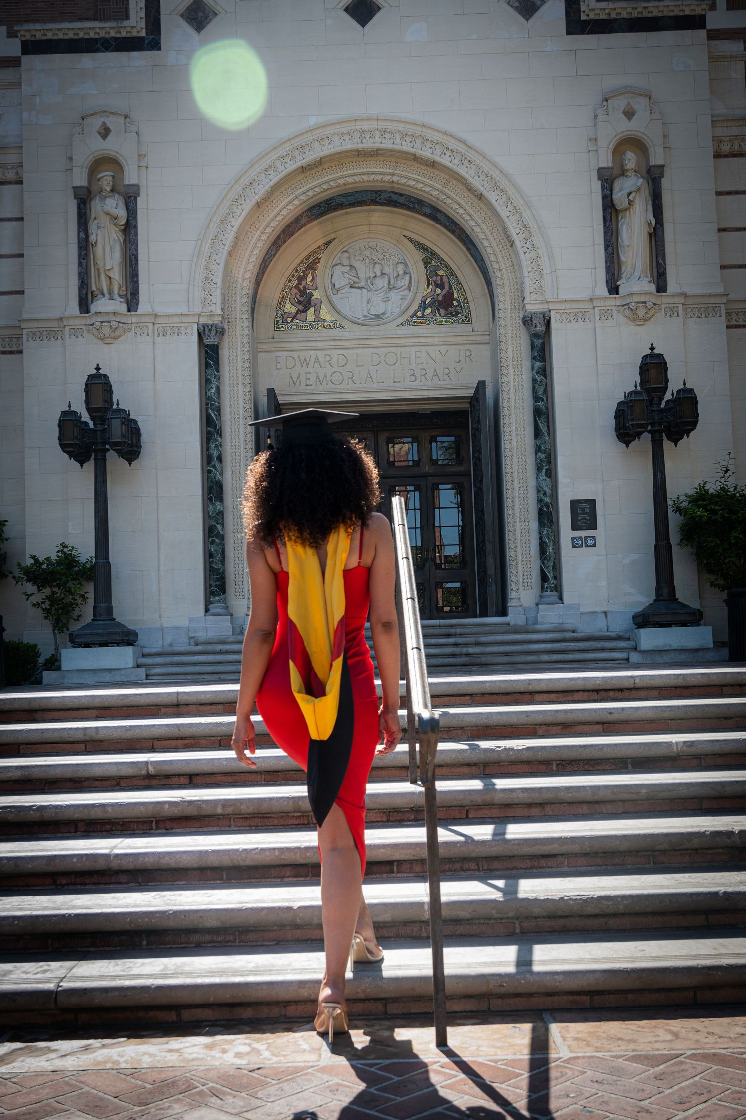 Graduate in red dress and gold stole ascending stone steps of ornate university building entrance.