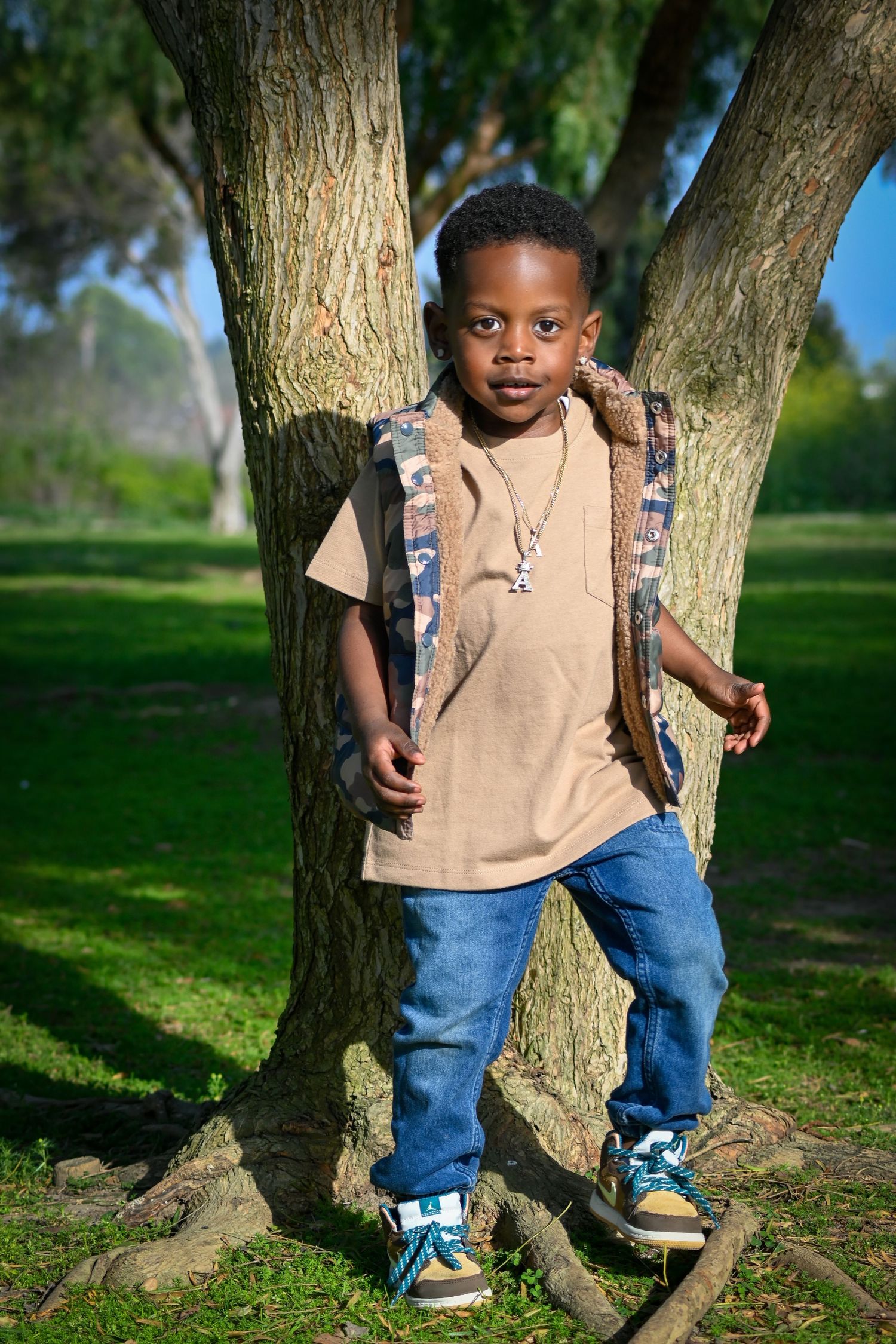 Young Black boy with backpack standing by tree roots in sunny park, wearing jeans and tan shirt.