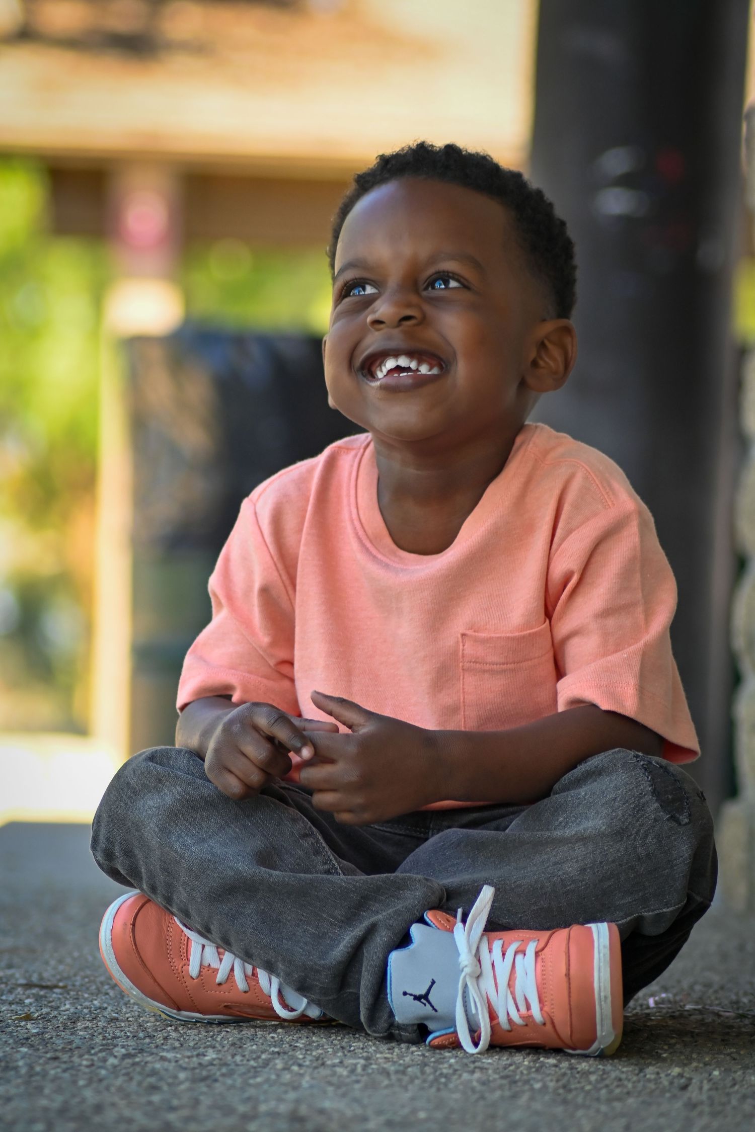 A happy young Black boy sitting cross-legged on the ground, smiling joyfully, wearing a pink shirt and orange sneakers.