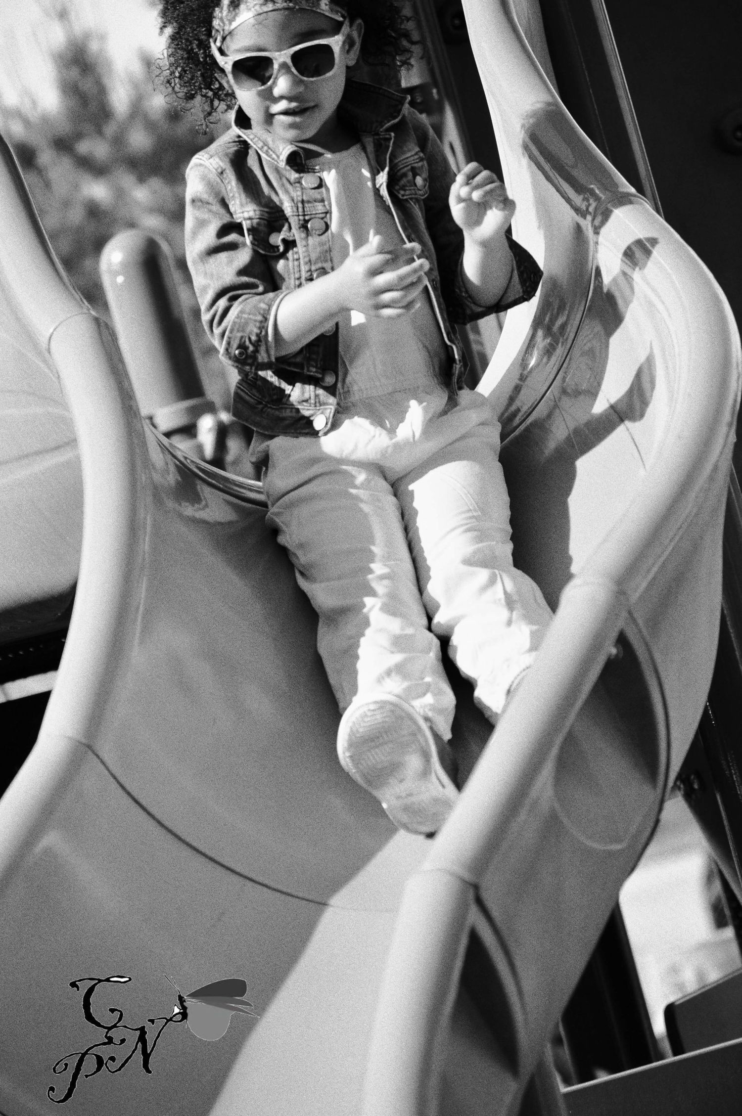 Black and white photo of a young girl wearing sunglasses sliding down a playground slide.