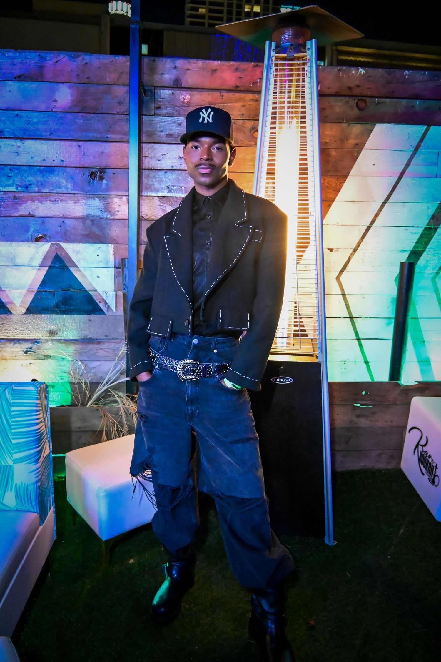 Someone wearing a NY Yankees cap, black leather jacket and baggy jeans poses in front of a colorfully lit wooden wall display.