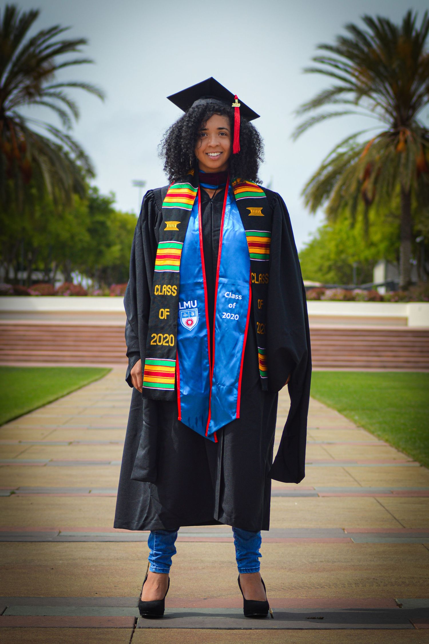 Graduate in cap and gown with colorful stoles stands on campus walkway with palm trees in background.