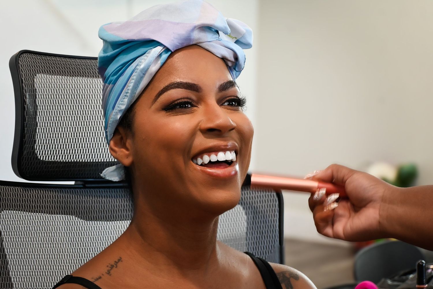 A woman with a blue head wrap smiles brightly while getting makeup applied in a salon chair.