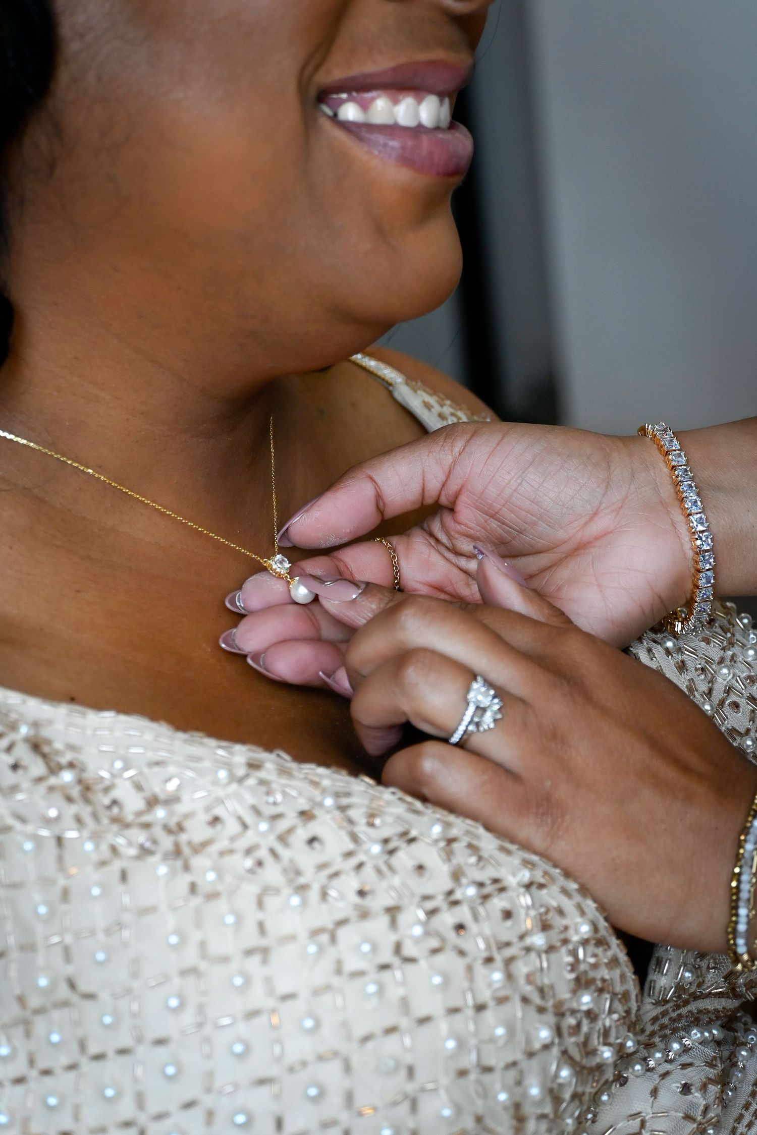 A bride smiles as someone clasps a delicate gold pearl necklace, wearing a diamond ring and bracelet with a beaded gown.