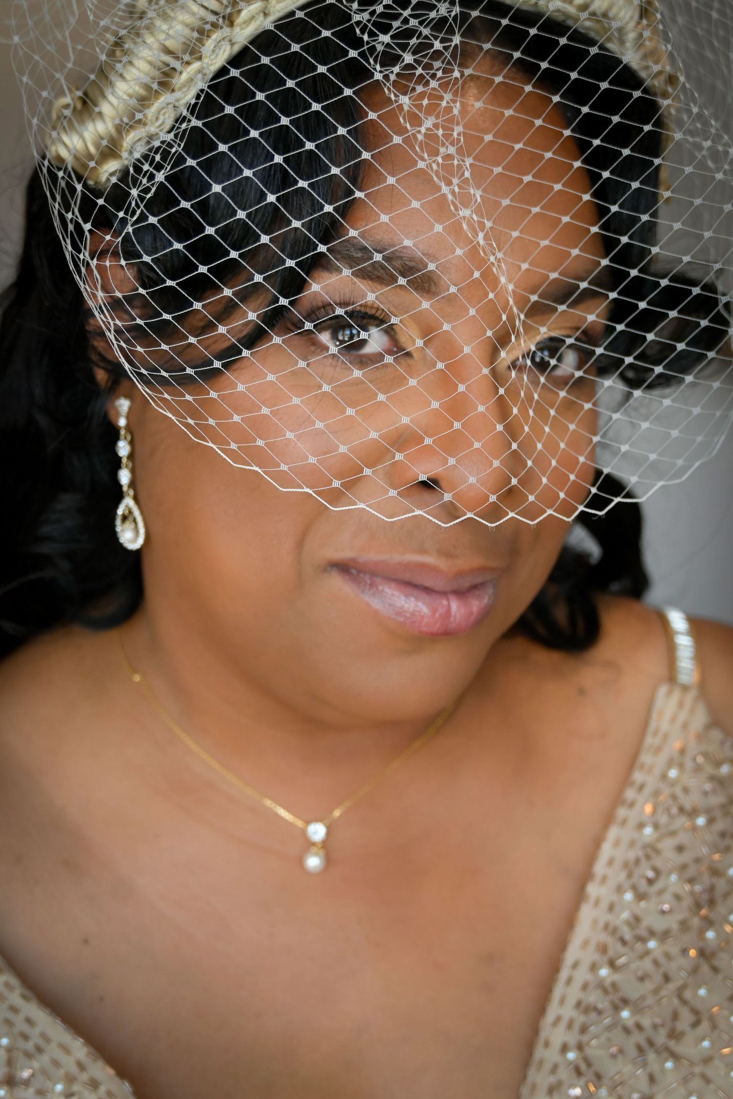 Elegant bride wearing a birdcage veil, pearl drop earrings, and gold necklace with soft bridal makeup.
