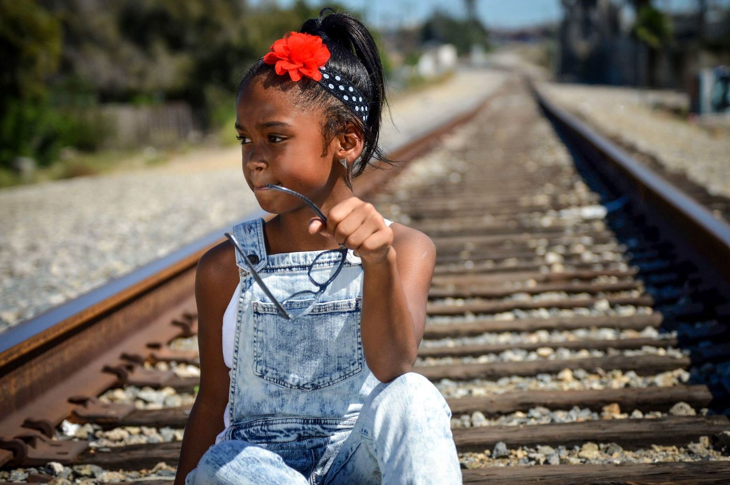 Young girl in denim overalls with red flower headband sitting on railroad tracks on a sunny day.