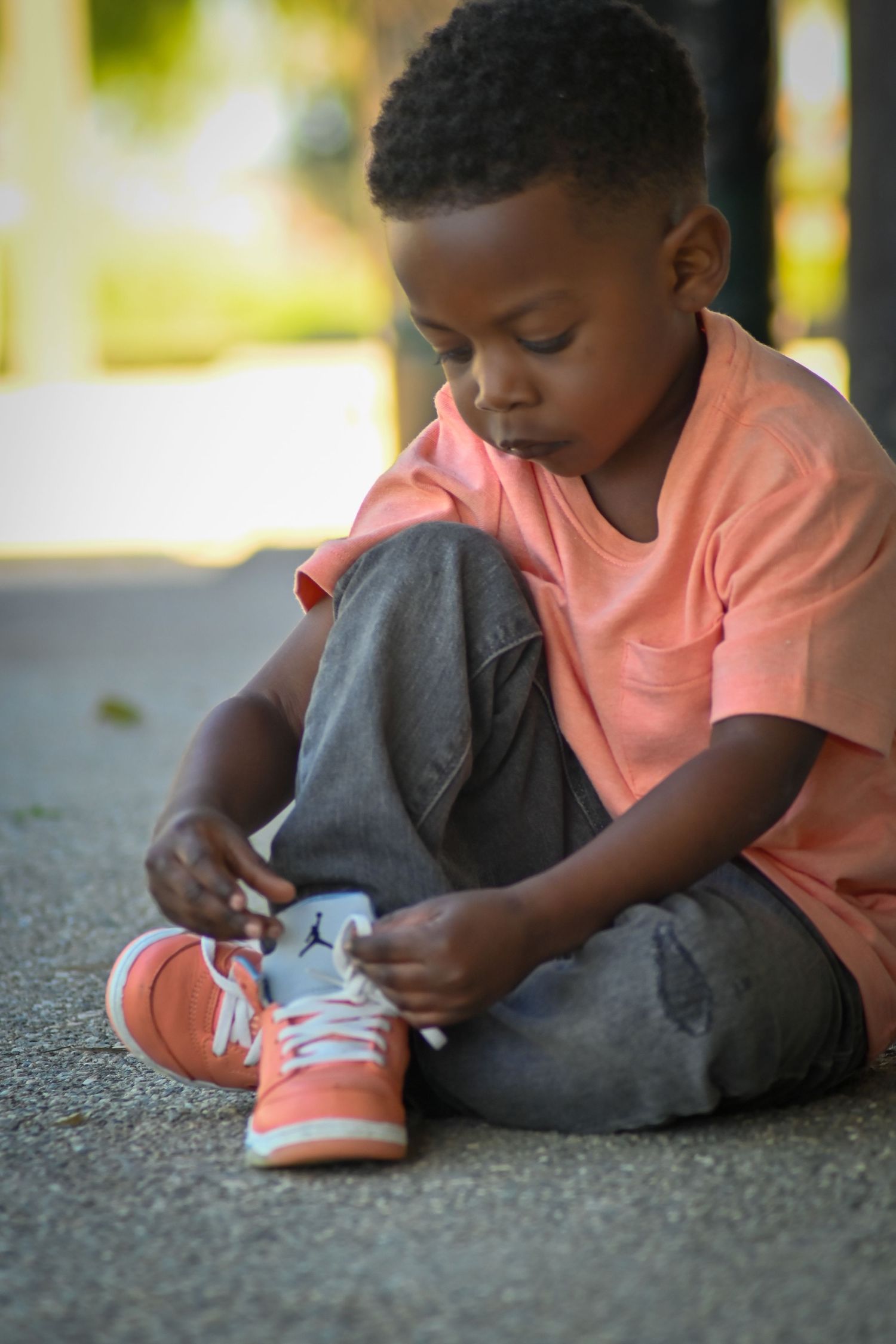 Young Black boy in orange shirt and jeans sitting on pavement tying his sneakers outdoors.