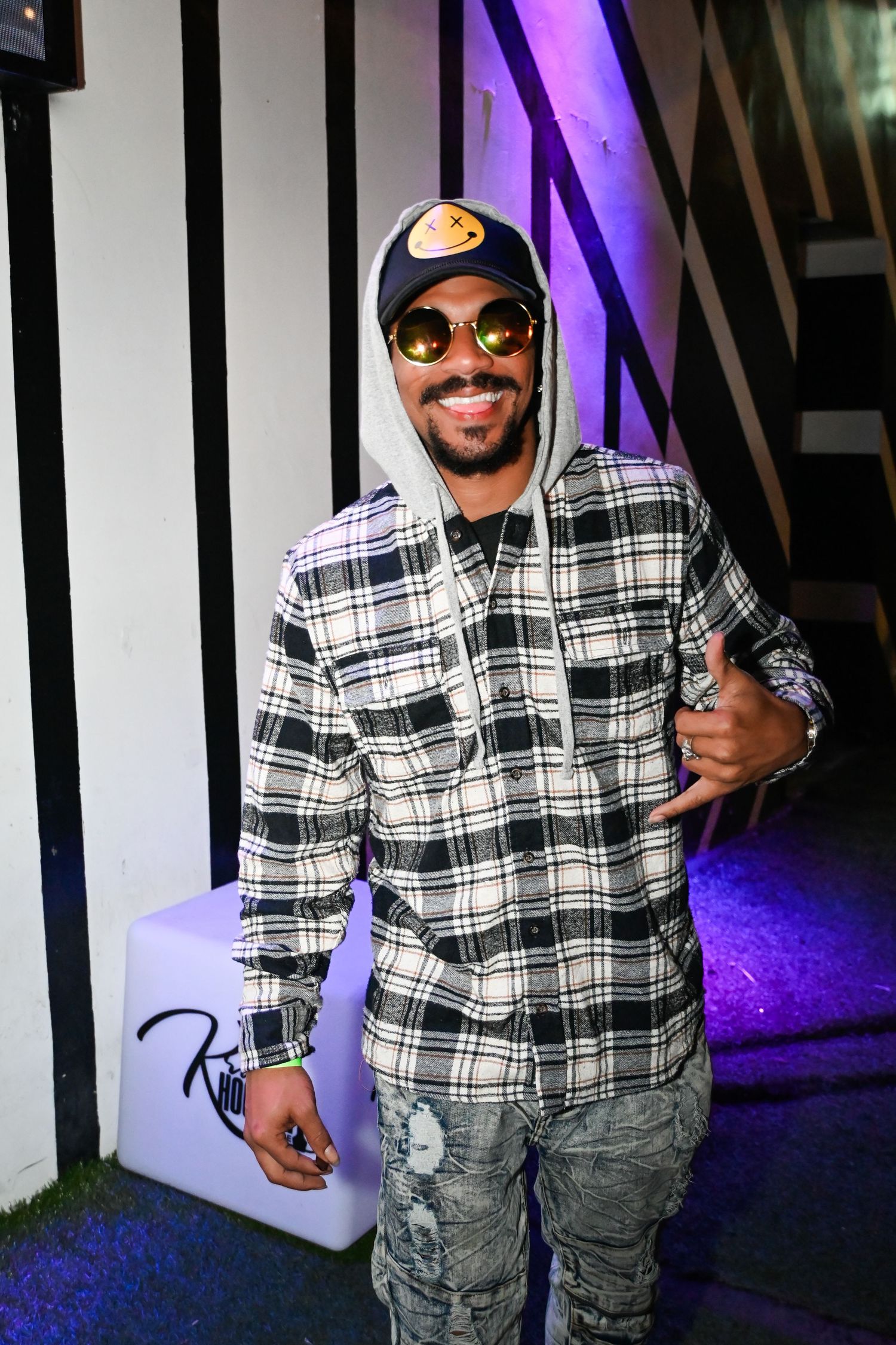 Someone wearing a black and white plaid shirt and gray cargo pants poses in front of purple lighting.