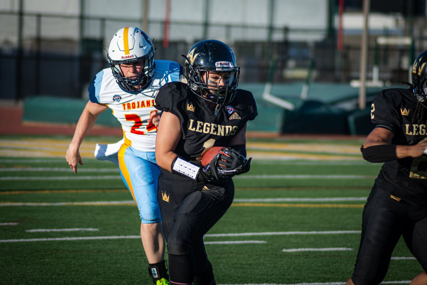 Football players in action, Legends player running past Trojans defender on a turf field during a game.