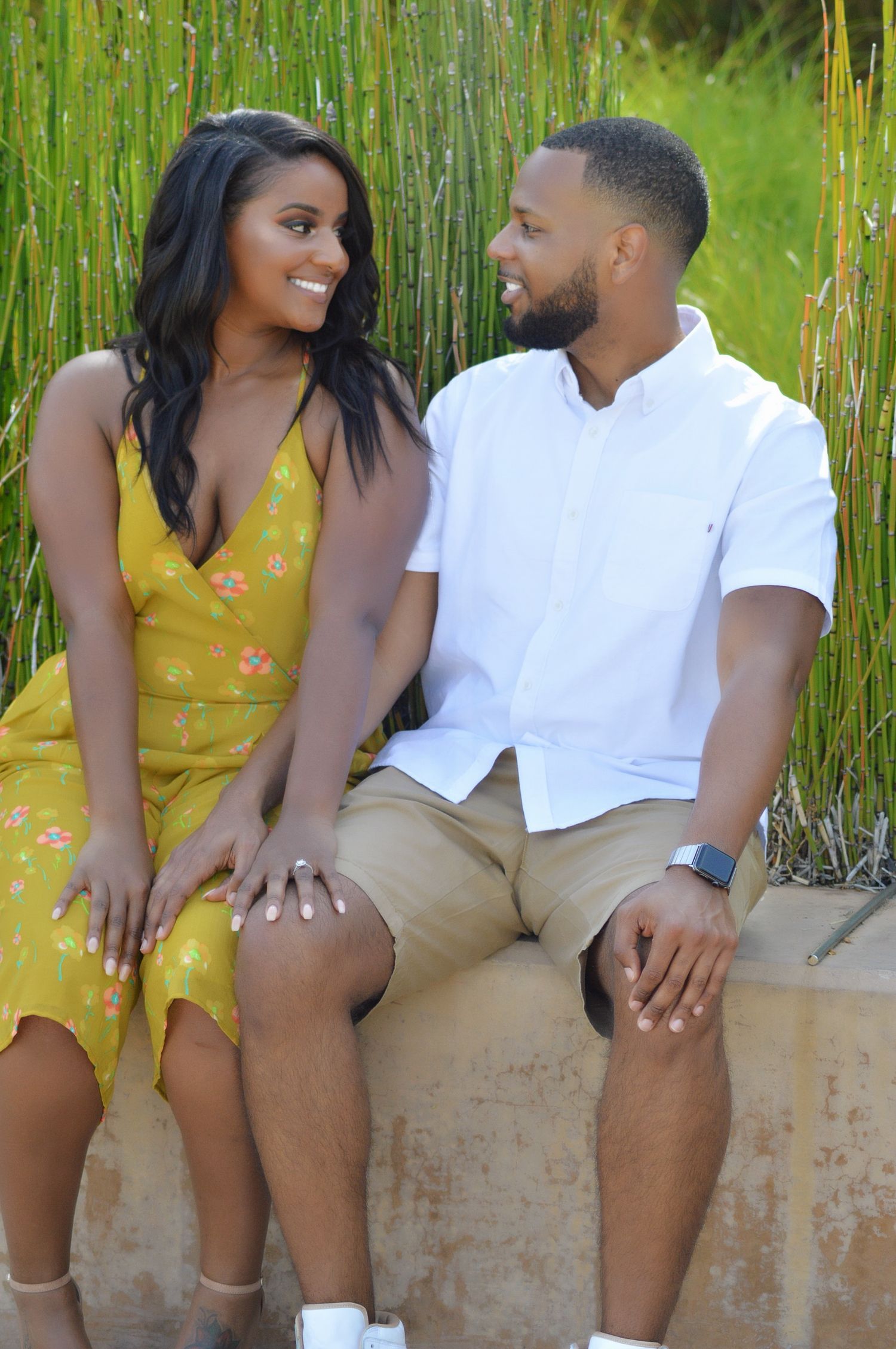 A smiling couple sits together outdoors, woman in yellow floral dress and man in white shirt, sharing a loving glance.