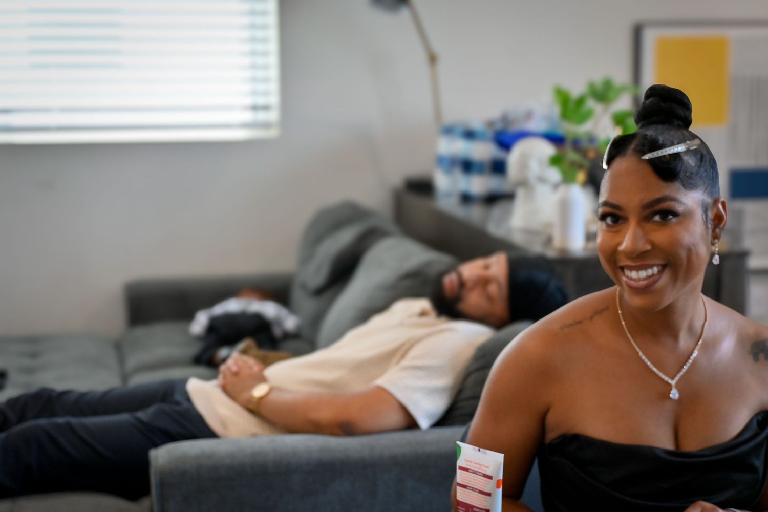 A smiling woman in a black top sits on a couch while a man relaxes in the background.