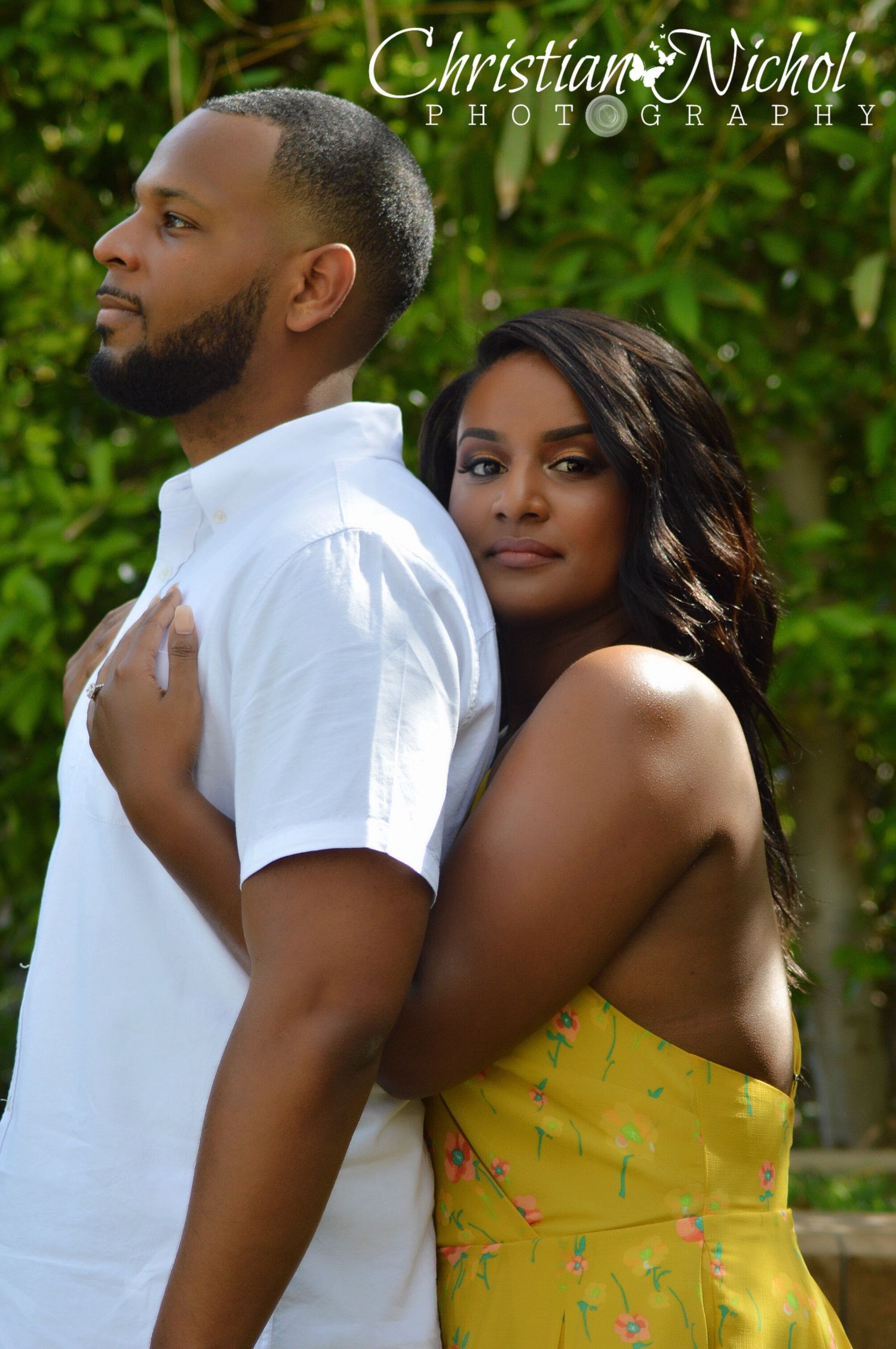 A couple poses back-to-back outdoors, man in white shirt, woman in yellow floral dress, photographed by Christian Nichol Photography.