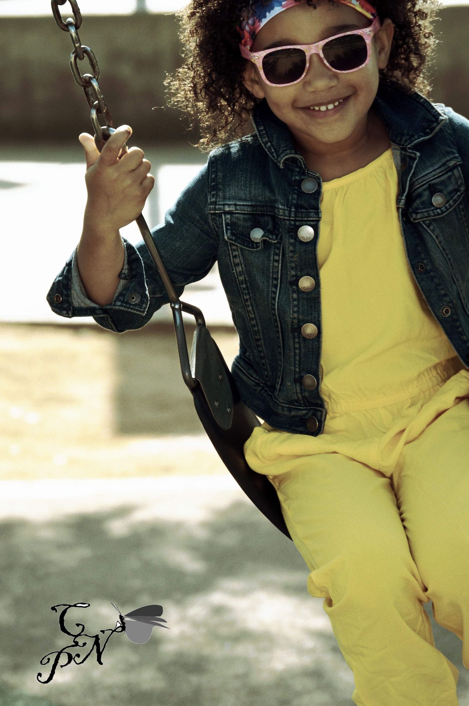 A smiling young girl wearing pink sunglasses, yellow outfit, and denim jacket sits on a swing at a playground.