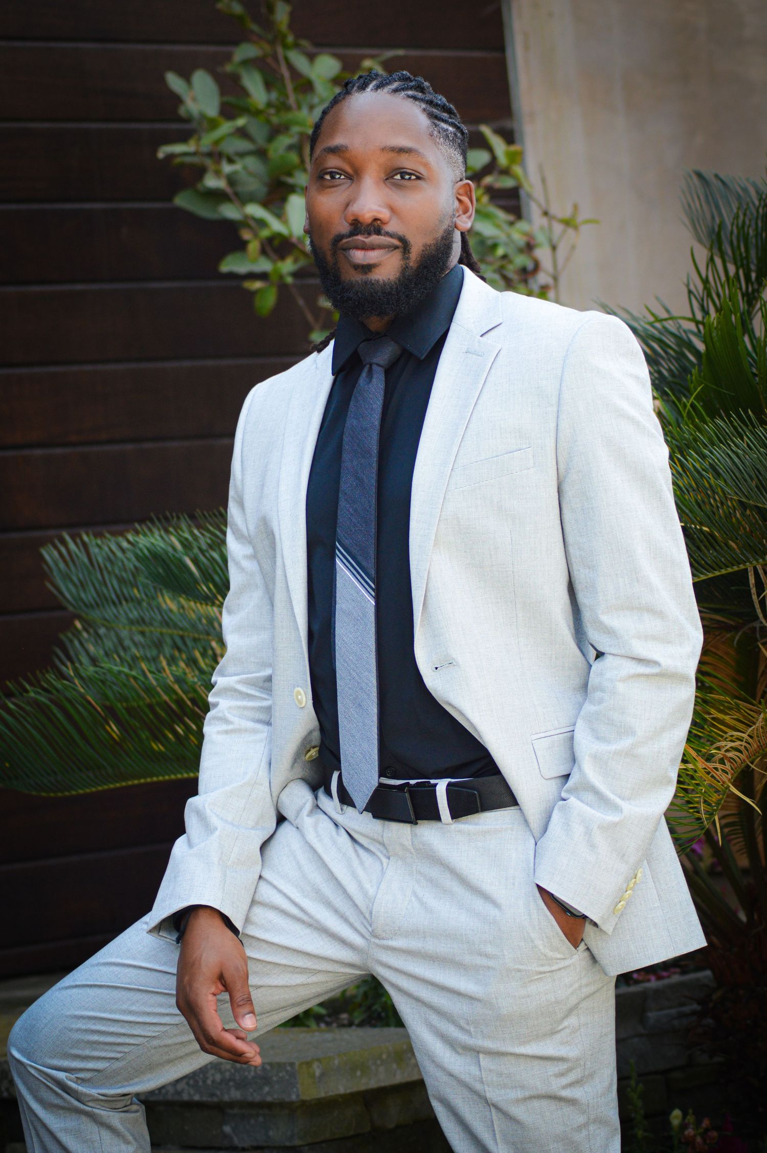 A person in a crisp white suit and black shirt poses outdoors against greenery in a series of professional portraits.