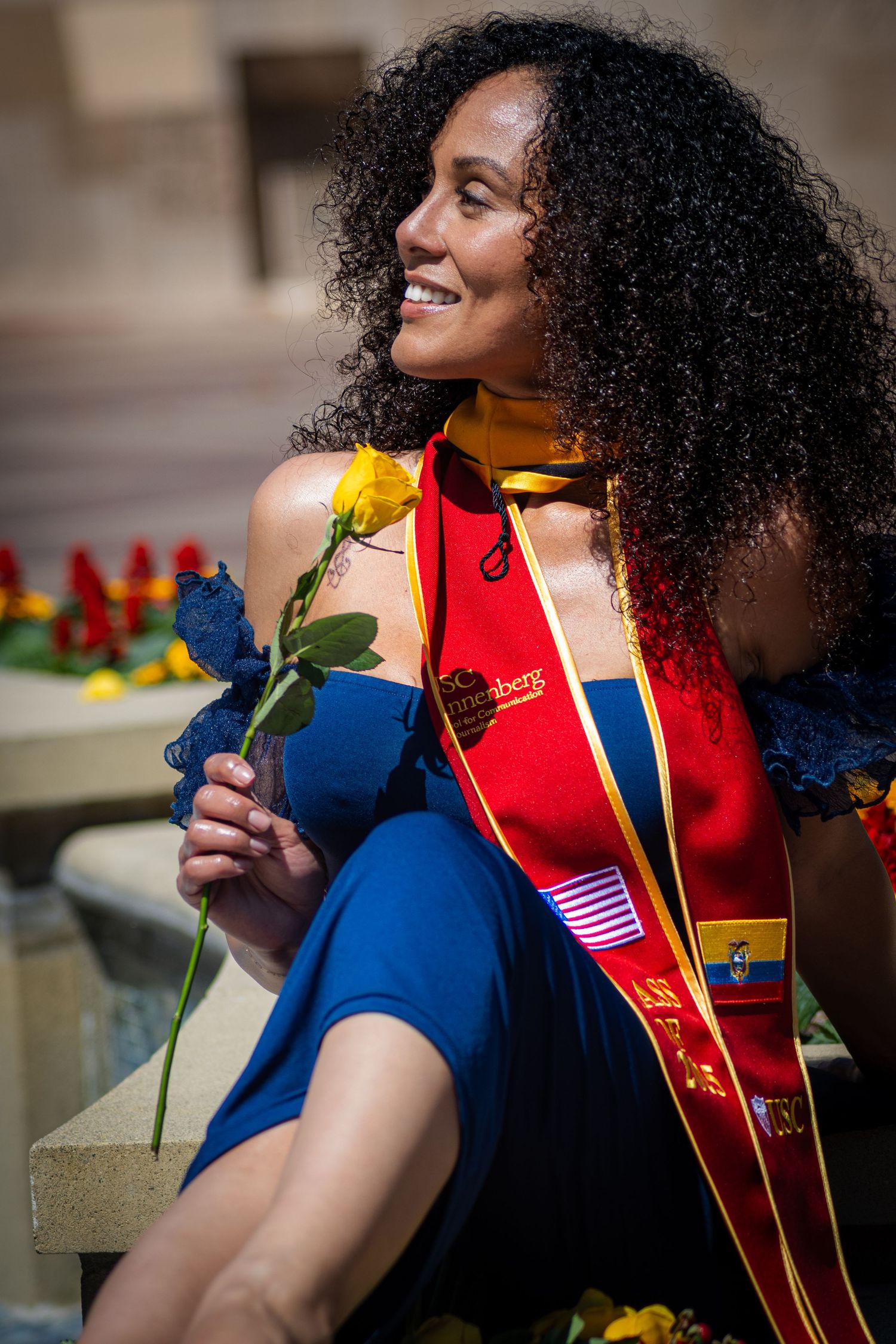 Graduate wearing stoles with national flags holds yellow flower in outdoor setting.