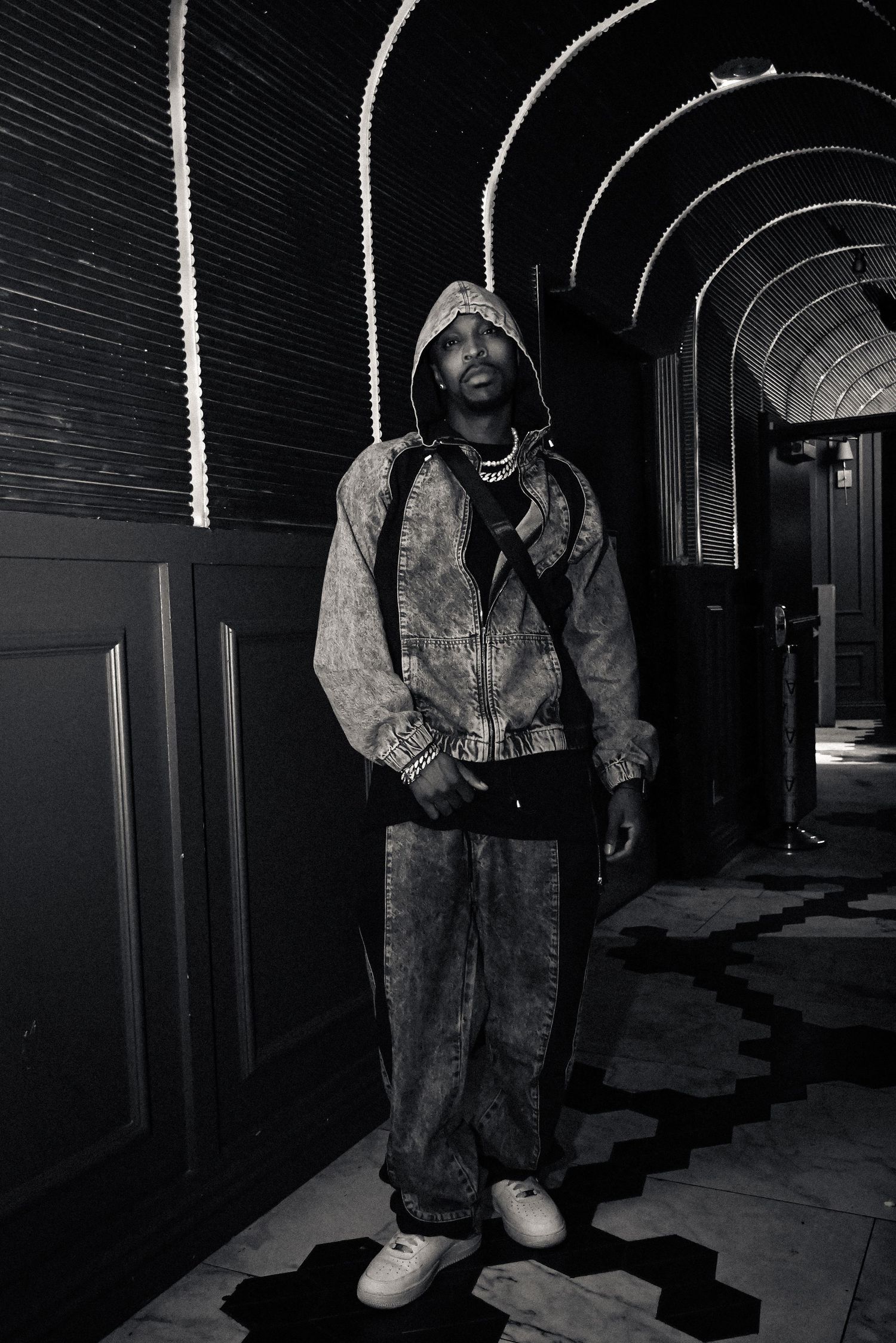 A person wearing a hooded jacket and baggy jeans stands in a dimly lit arched hallway with dramatic lighting.