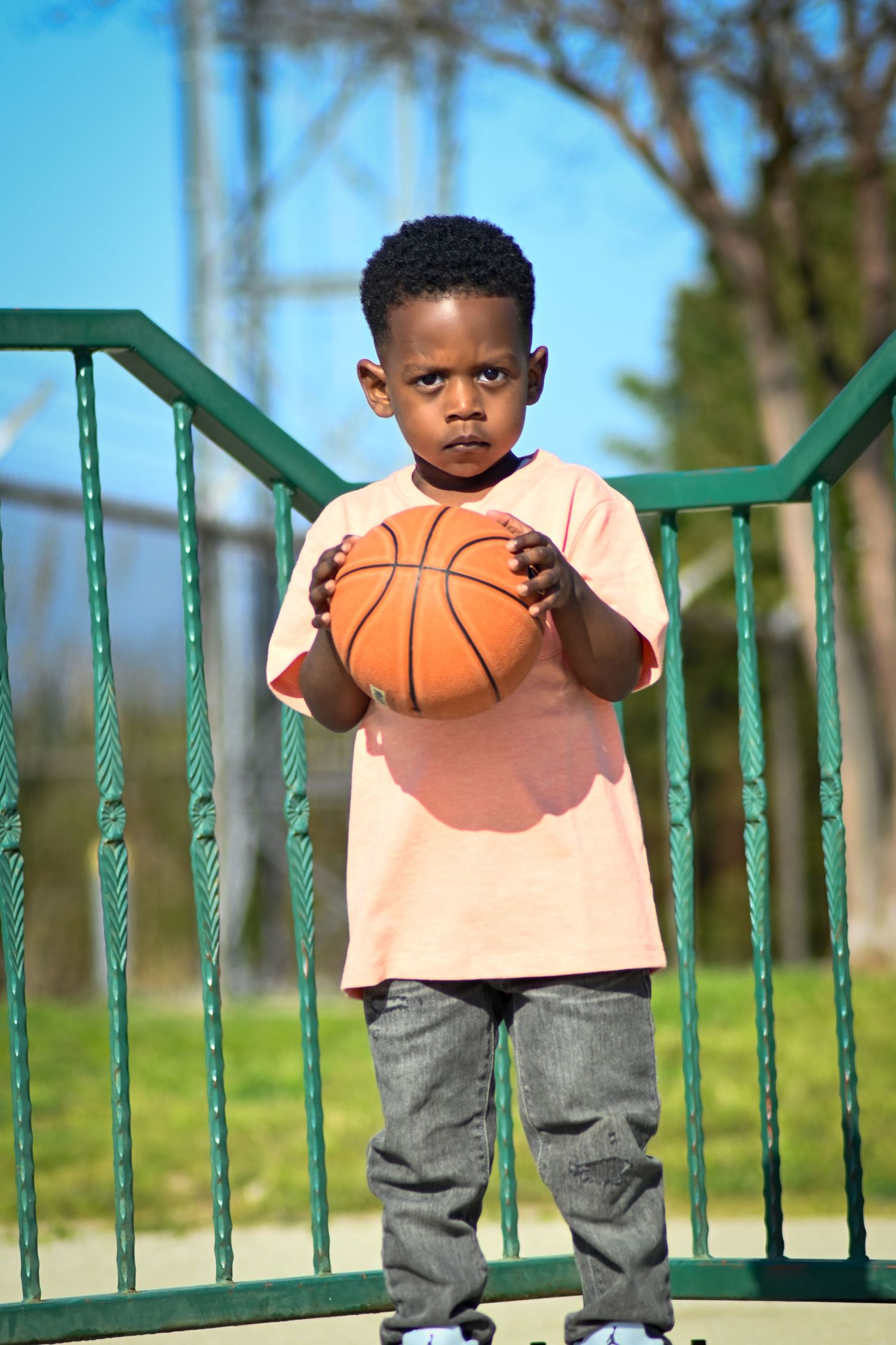 Young boy holding a basketball outdoors on a sunny day, standing near green railings on a playground.
