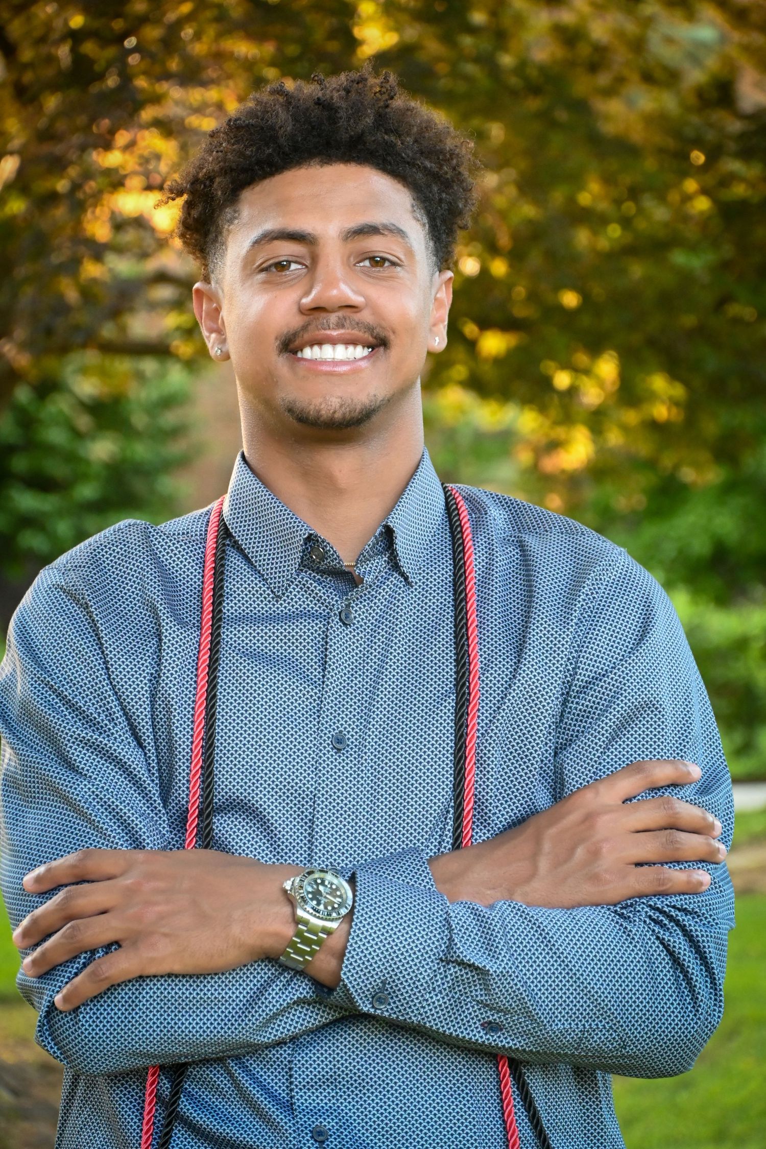 Professional portrait outdoors showing a confident smile, blue button-down shirt, and red suspenders against autumn foliage.