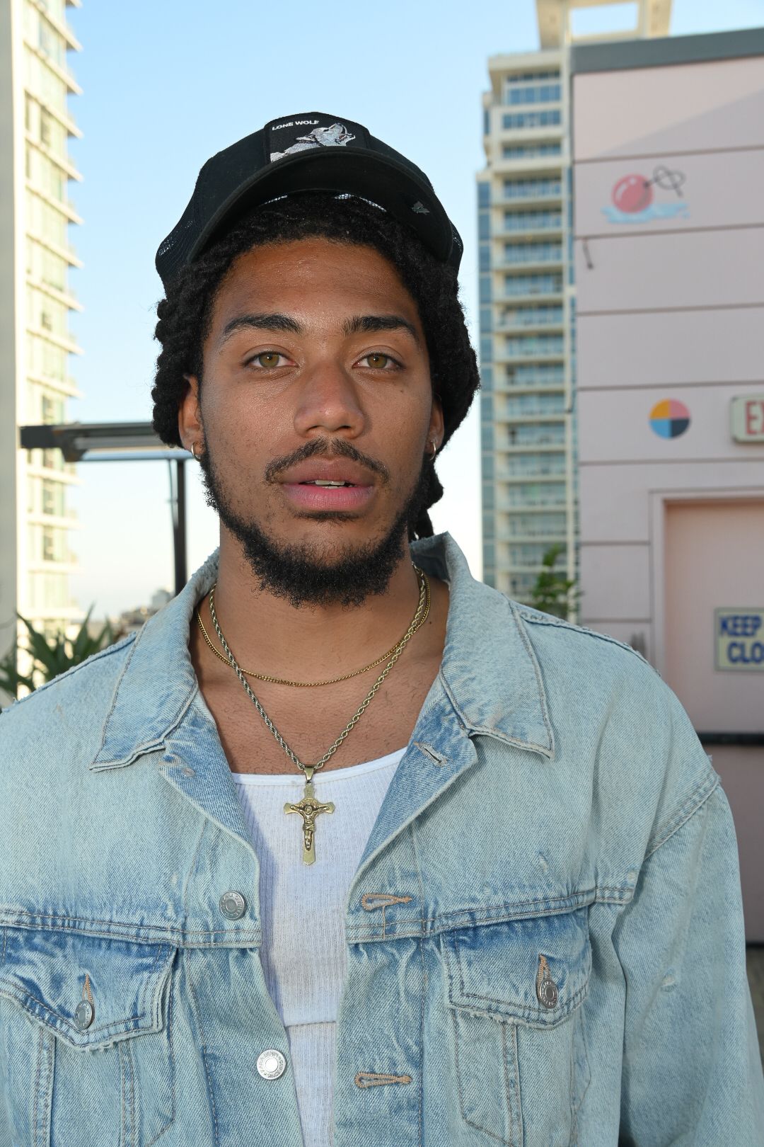 A person wearing a light denim jacket and black beanie poses against a building exterior.