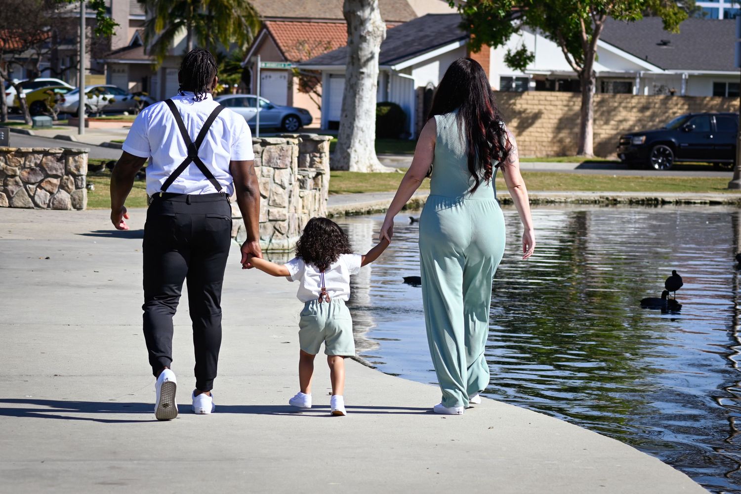 People walking along a waterfront path with ducks swimming in the water on a sunny day.