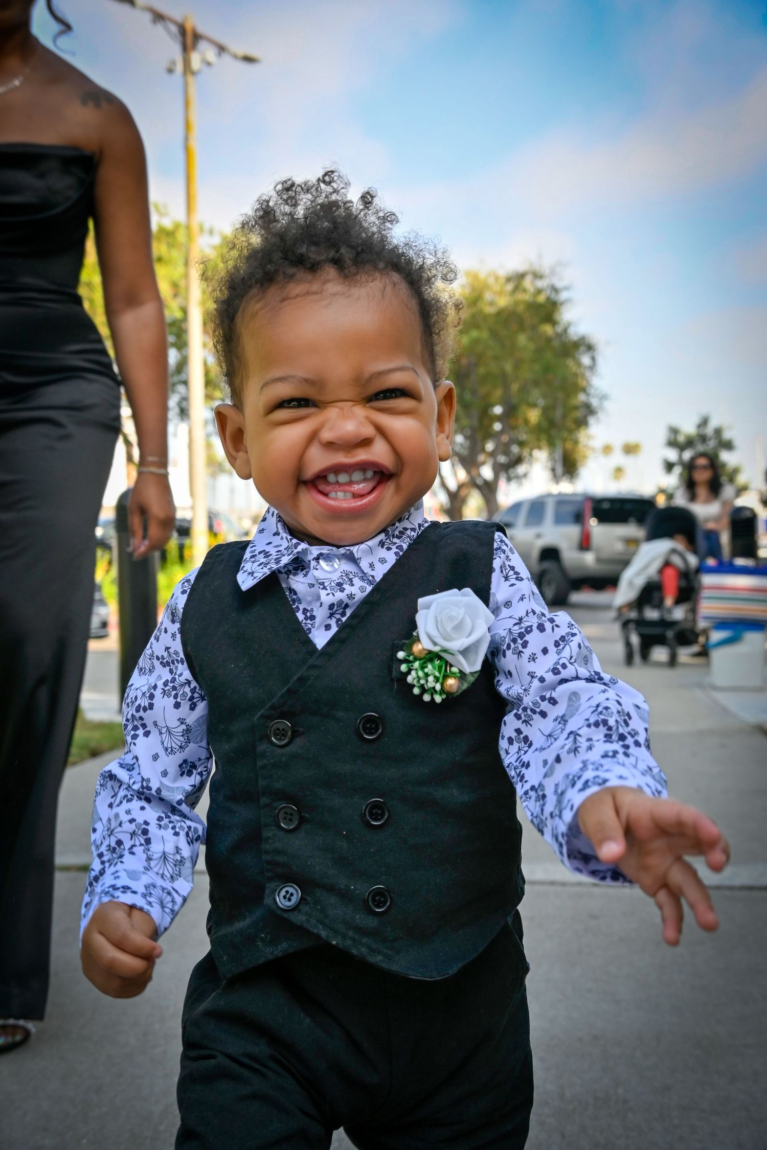 Adorable smiling toddler boy dressed in a black vest and floral shirt with a white rose boutonniere at an outdoor event.