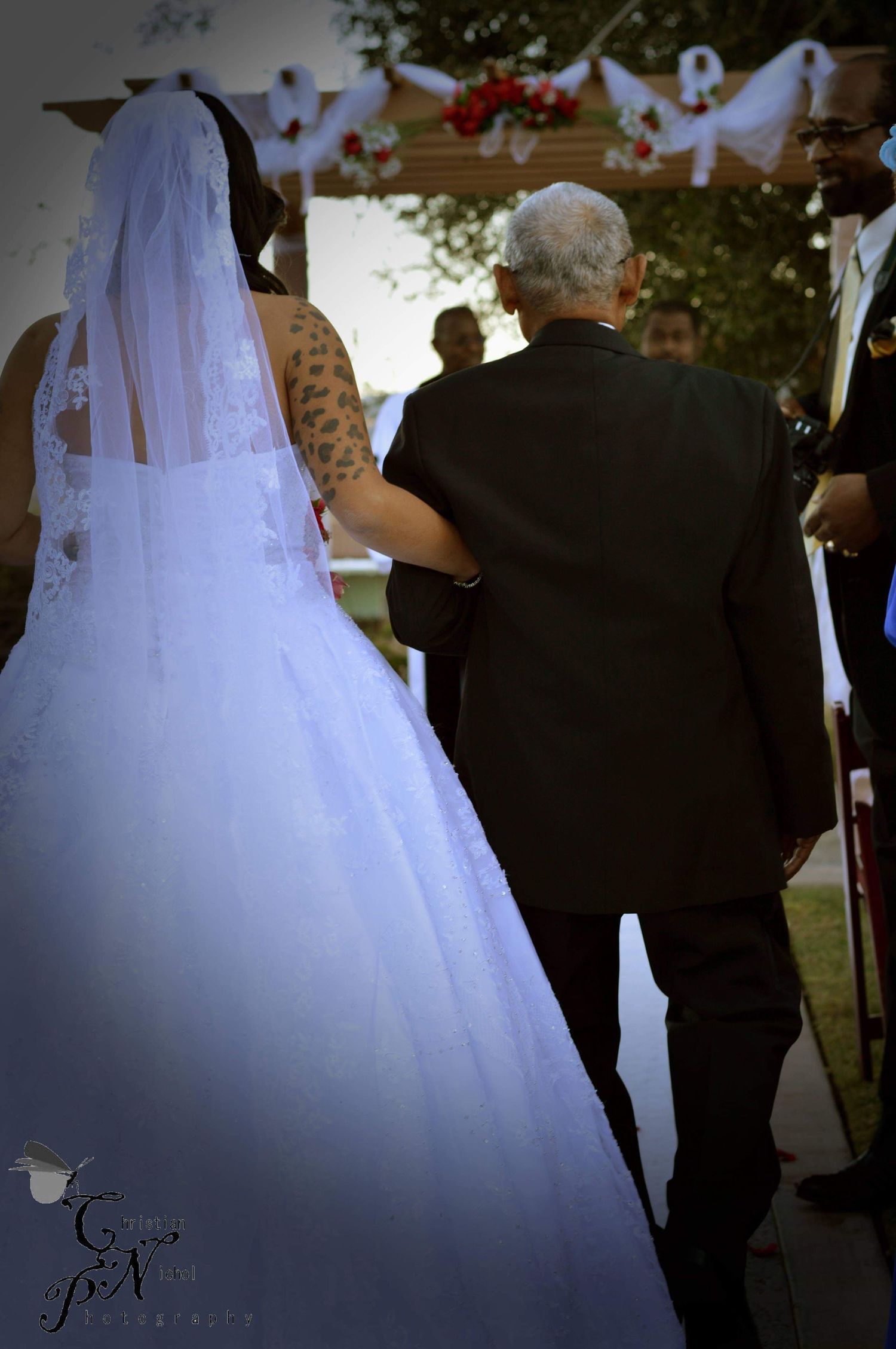 Bride in white ball gown and veil walks down the aisle with escort at outdoor wedding ceremony.