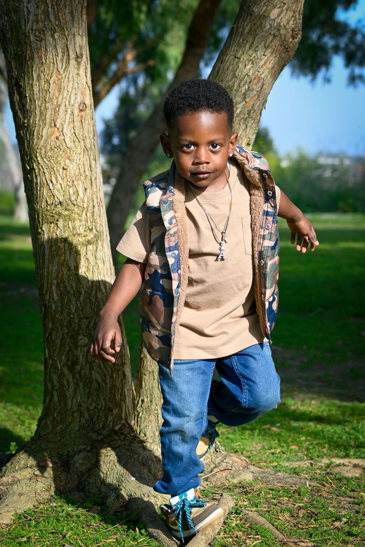 Young Black boy in jeans and vest leans against a tree trunk in a sunny green park, wearing a necklace.