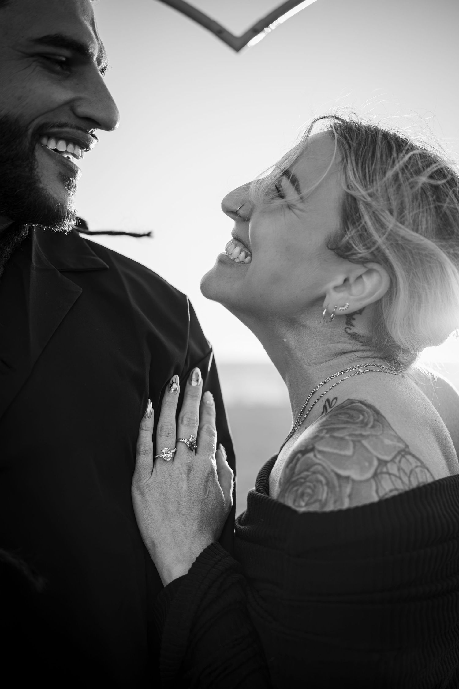 Black and white photo of a laughing couple, woman with tattoos and engagement ring, sharing a joyful moment outdoors.