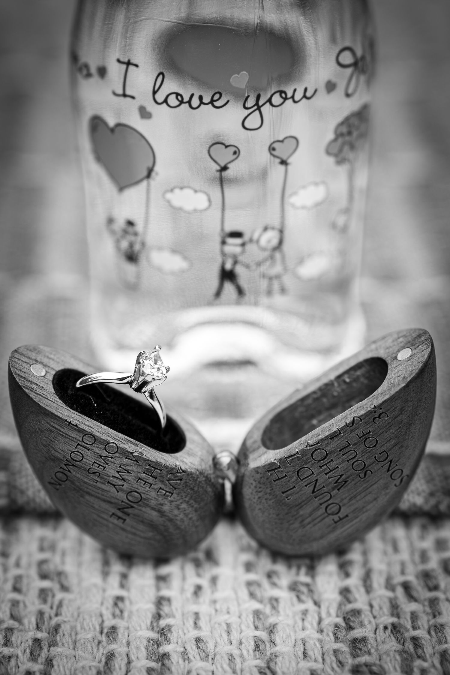 Black and white photo of a diamond engagement ring in a wooden heart-shaped box with an I love you glass jar in background.