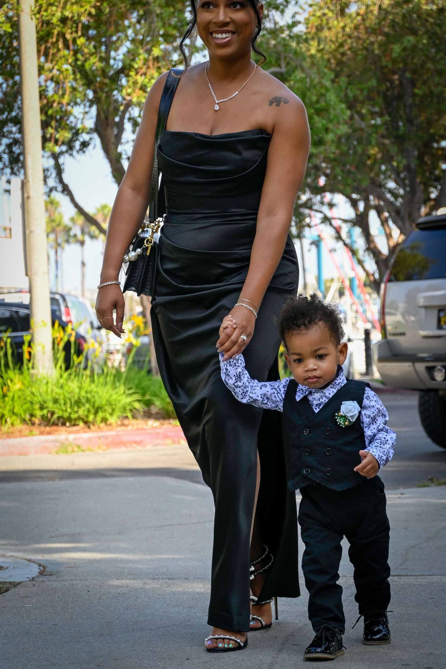 Woman in black gown walks hand-in-hand with toddler boy in suit on sidewalk at formal event.