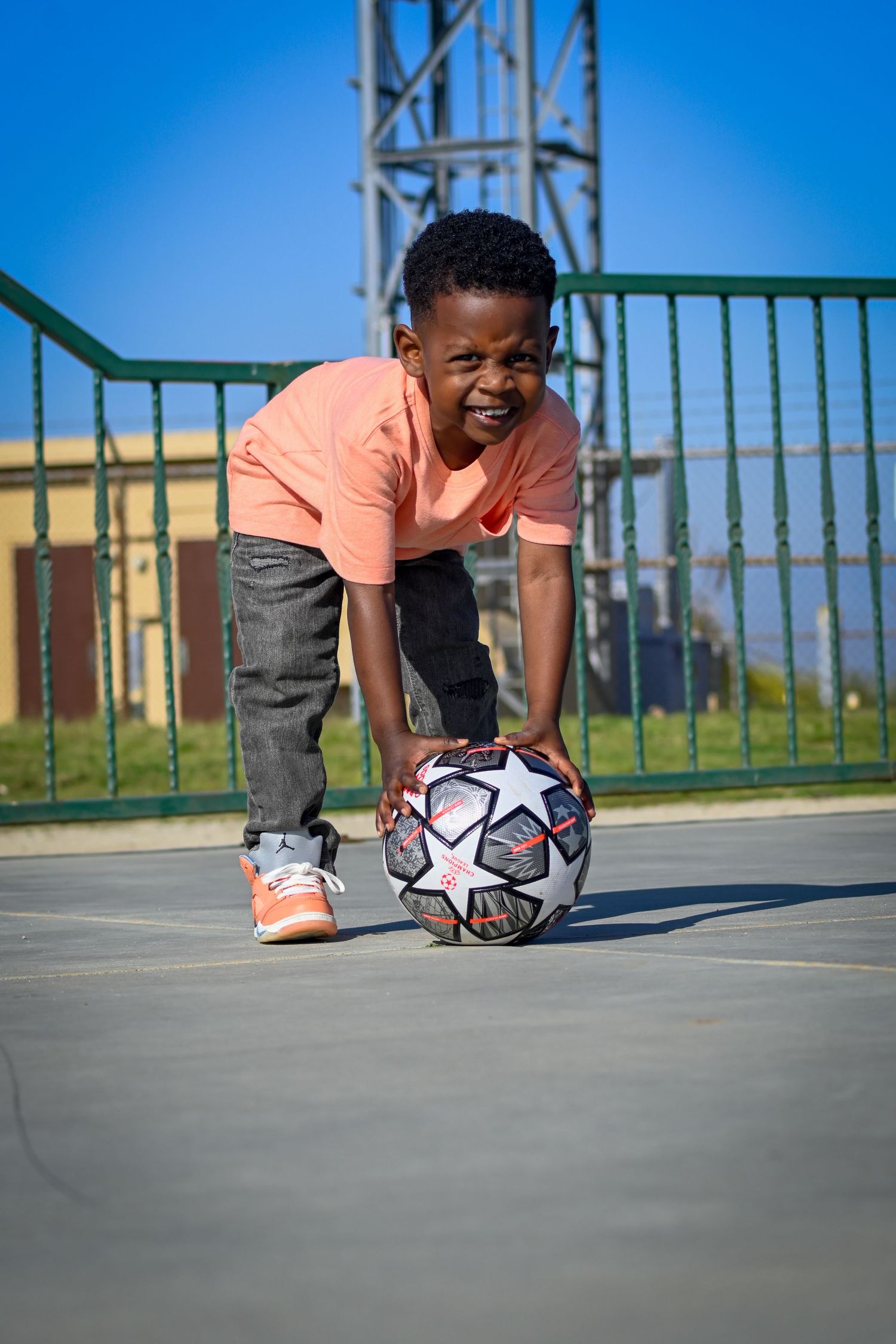 Young boy in orange shirt smiling while holding a soccer ball on an outdoor court on a sunny day.
