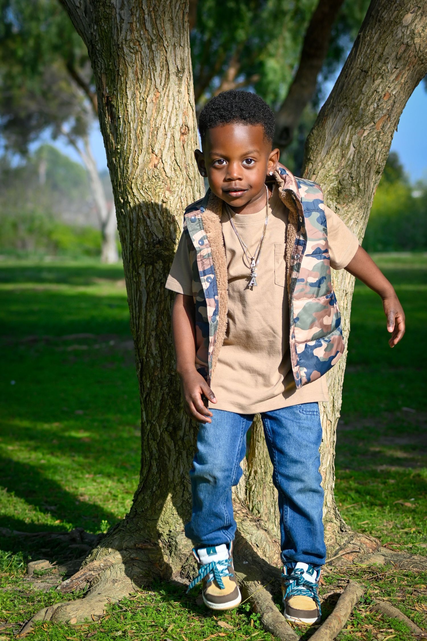 Young Black boy in camo vest and jeans poses confidently by a tree in a sunny green park.