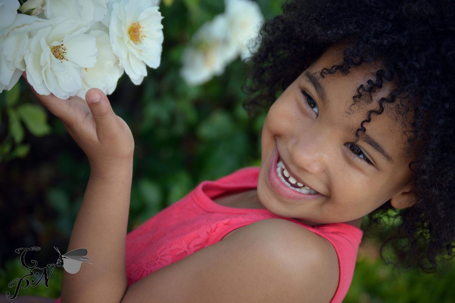 Smiling young girl with curly hair holding white roses, wearing a pink top, outdoors.