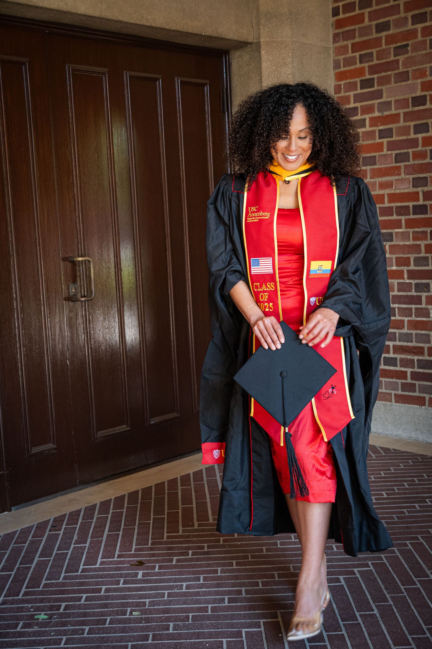 Graduate in cap and gown holds graduation cap while posing near wooden door.