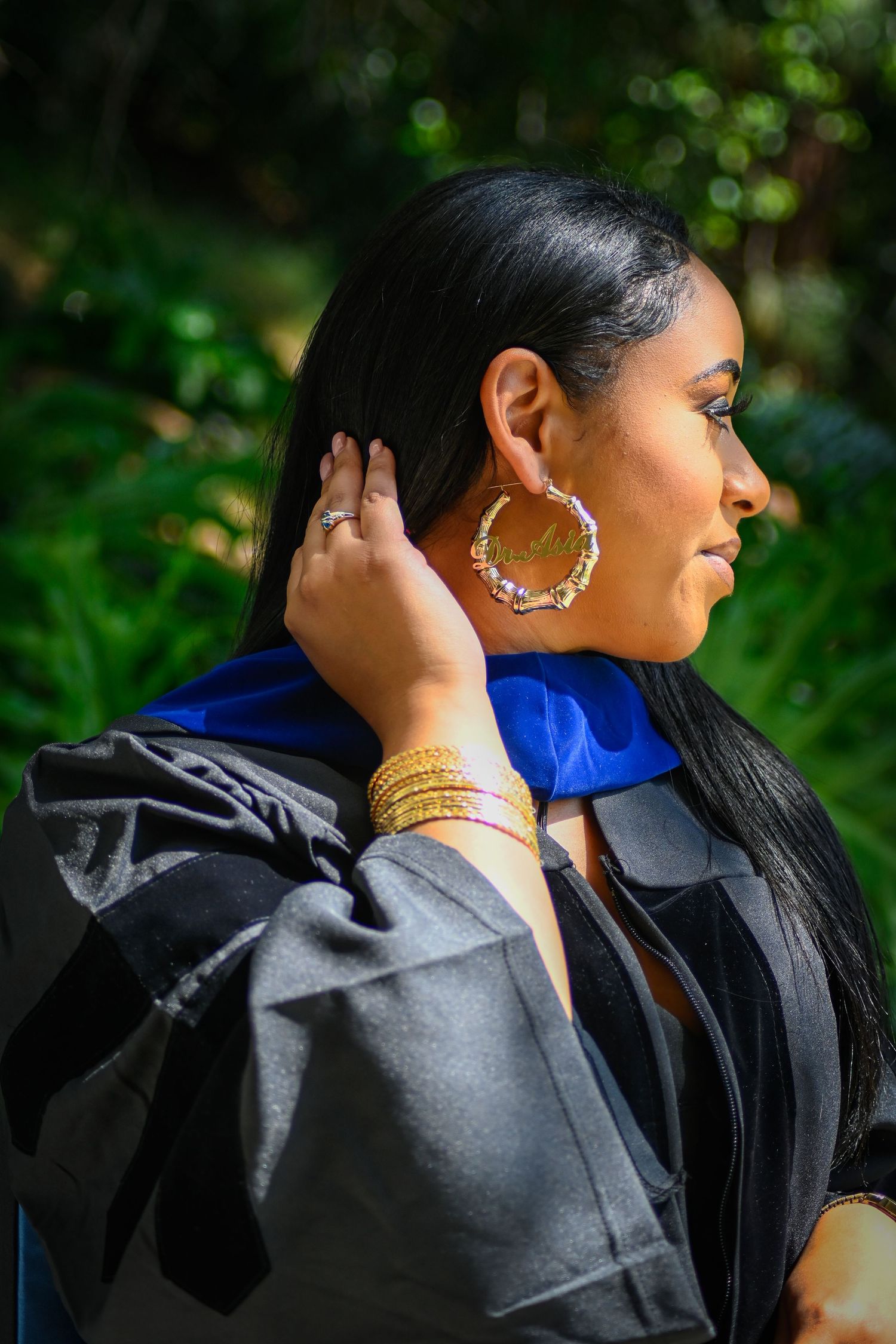 Close-up of graduation attire with gold jewelry and blue stole against natural green background.