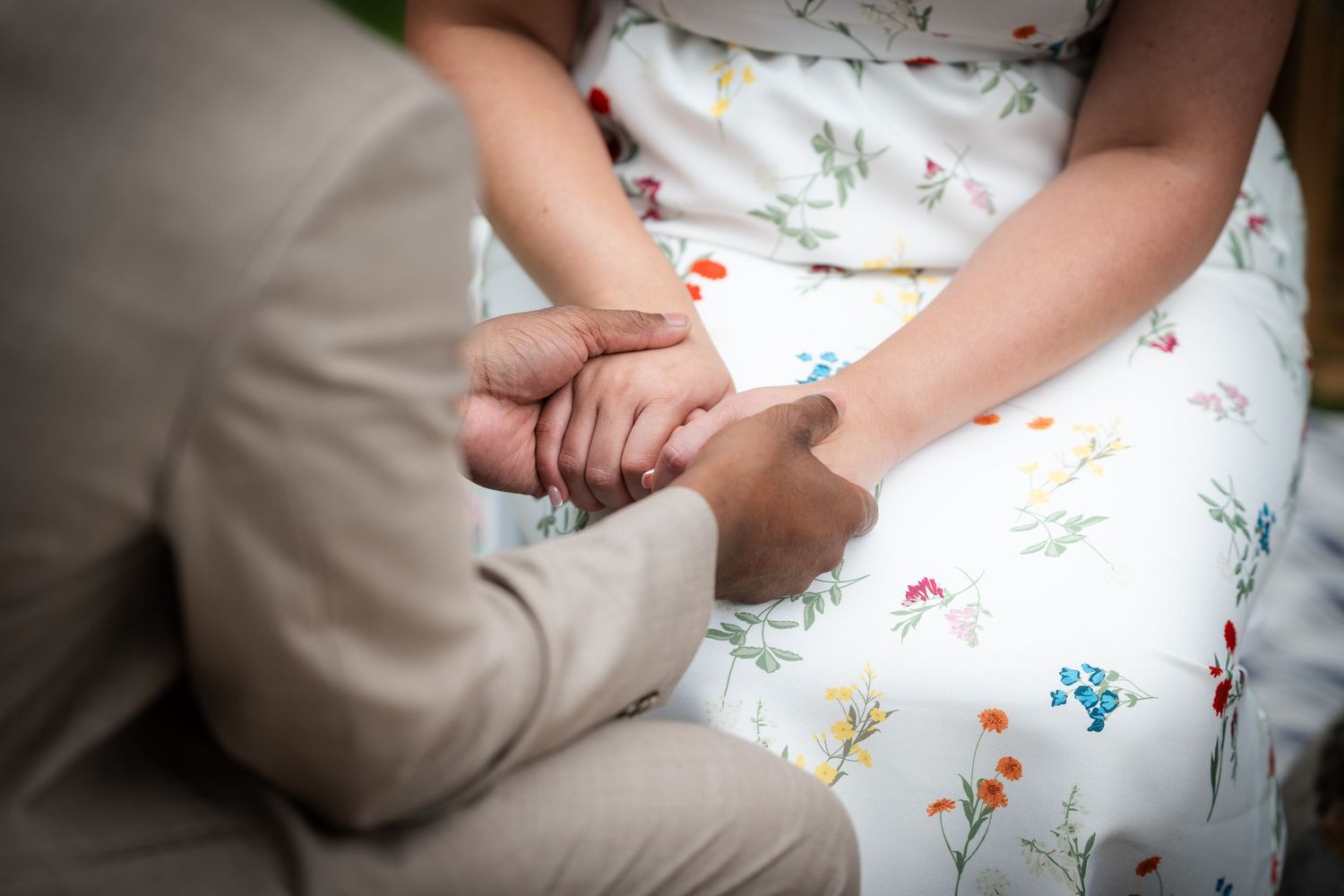 Couple holding hands tenderly, woman wearing a white floral dress during a romantic outdoor moment.