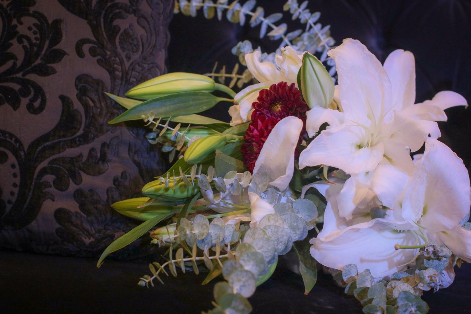 White lilies and red flowers bouquet with eucalyptus on dark floral sofa.