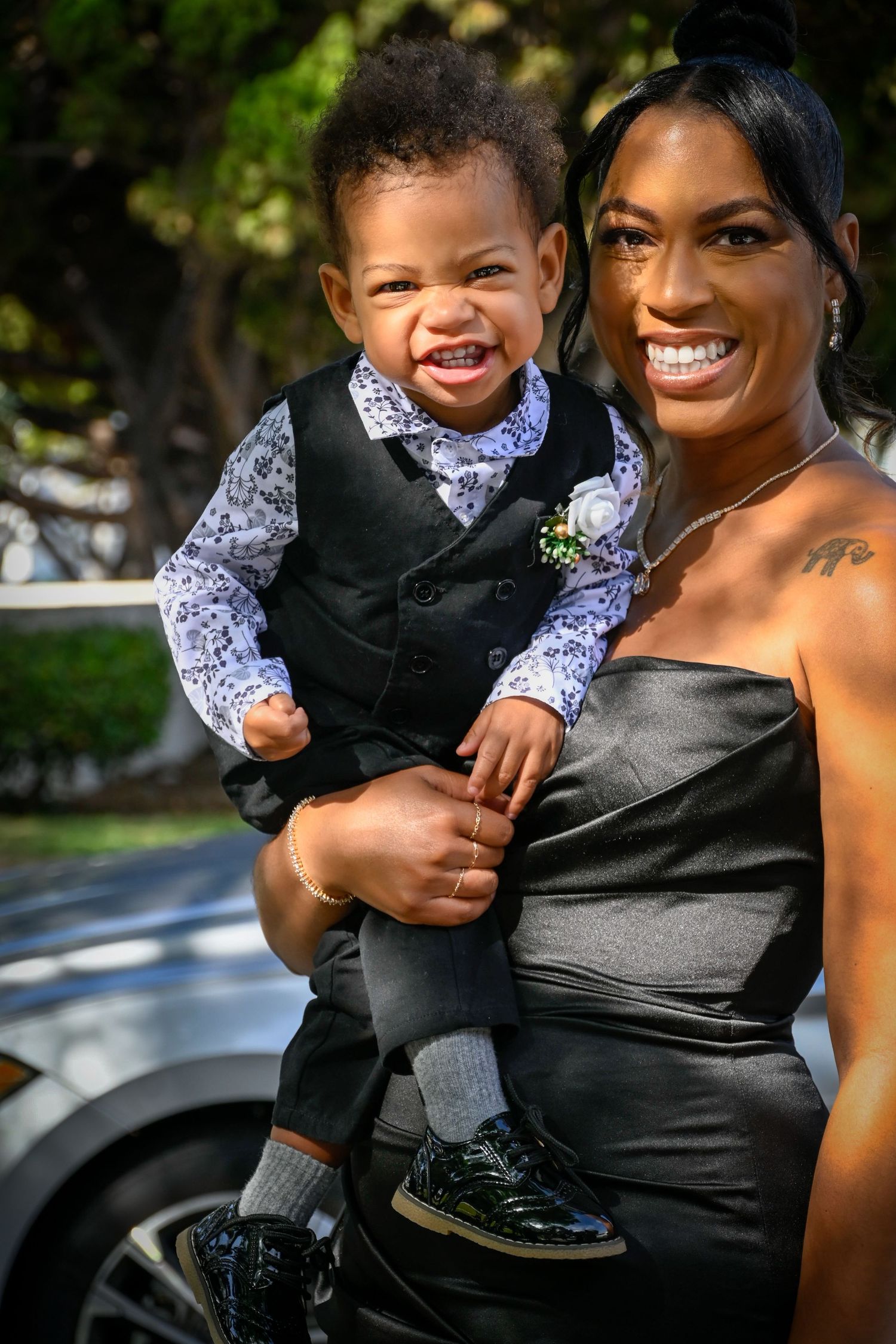 A smiling woman in a black strapless gown holds a laughing toddler boy dressed in a dapper black vest and floral shirt.