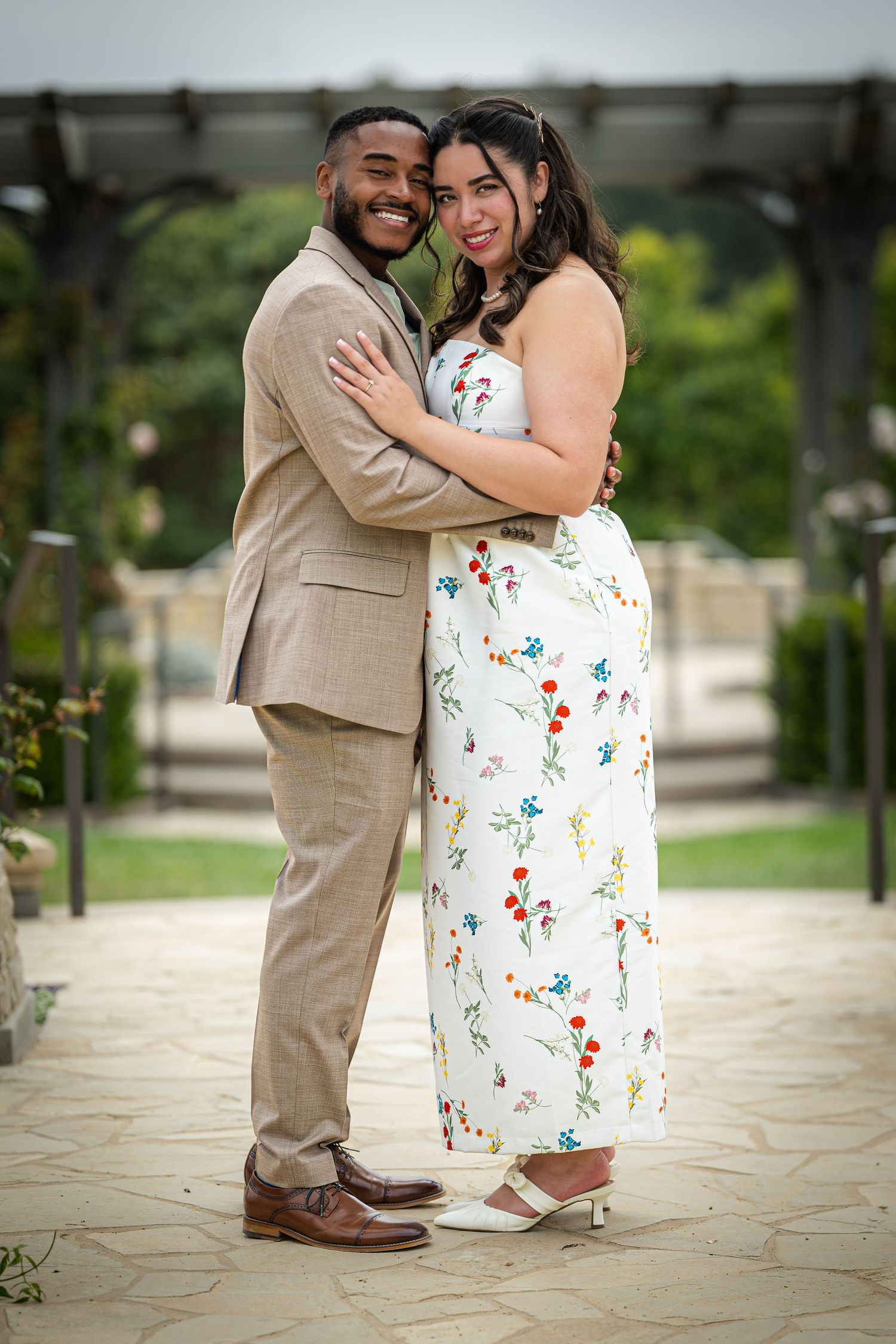 Couple embracing outdoors, man in tan suit and woman in white floral dress, posing in a garden setting.