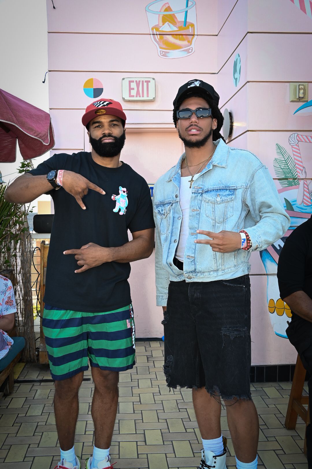 Two people pose together in casual outfits including black shirts, striped shorts and denim jacket against pink wall.