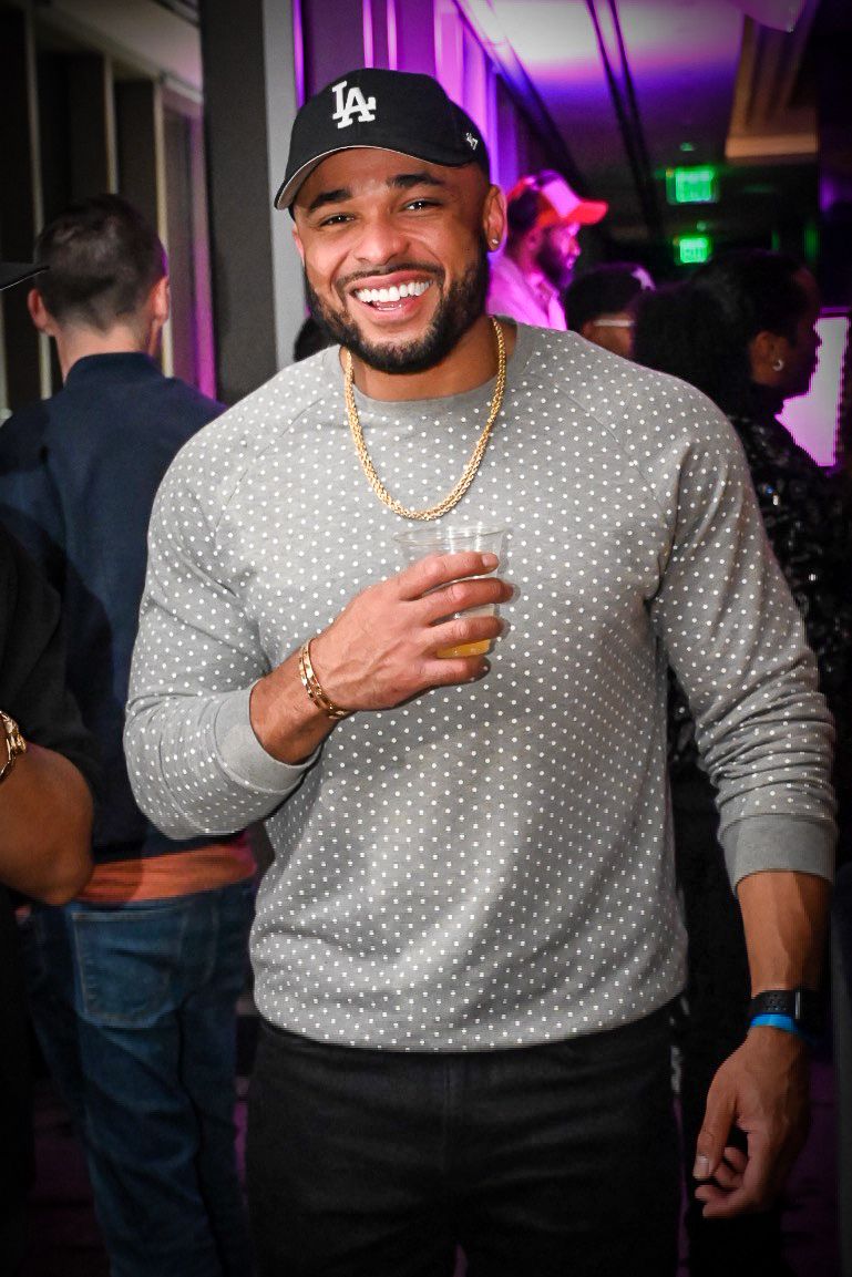 Person in gray polka dot sweater and LA baseball cap smiling at nightclub with purple lighting in background.