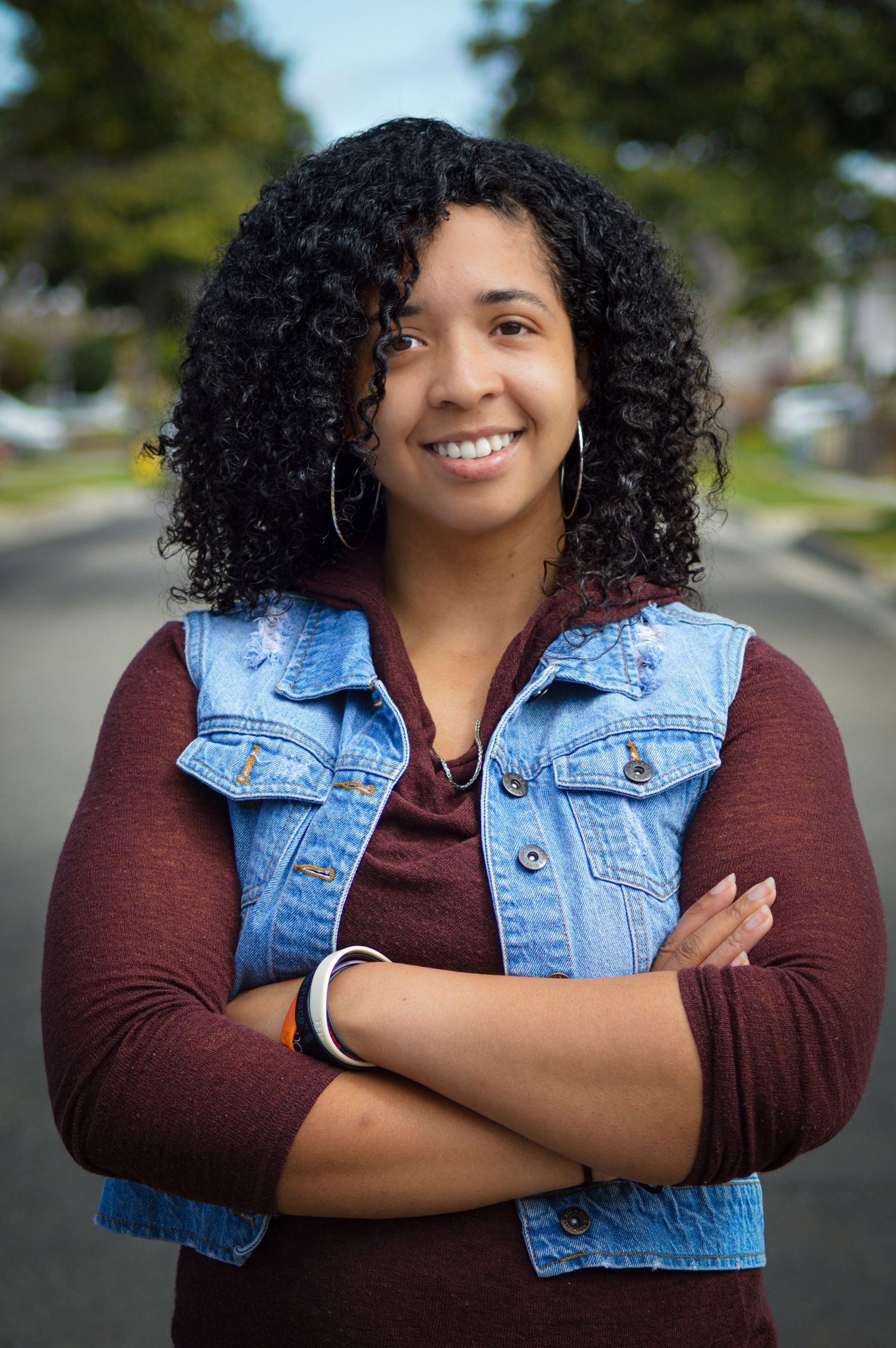 A person with curly hair smiles confidently while wearing a denim vest and burgundy top on a suburban street.