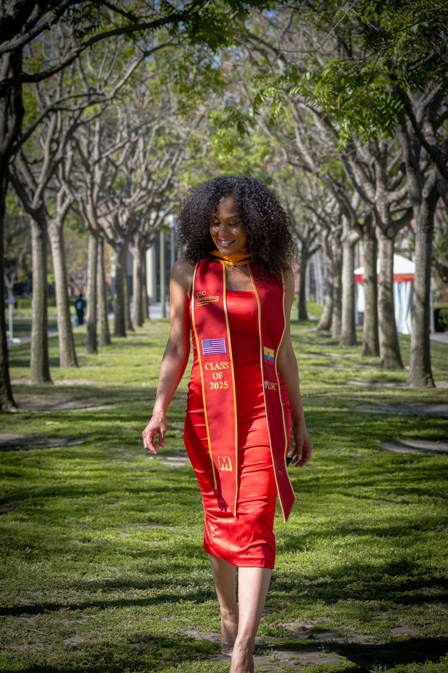 Graduate in red dress walks through tree-lined path wearing graduation stole.