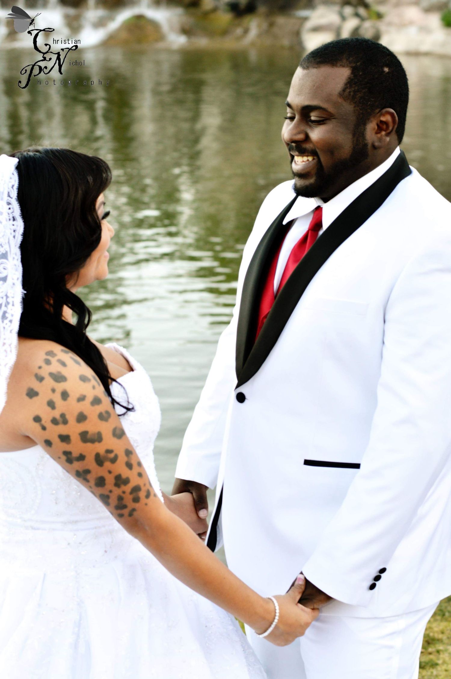 Interracial couple holding hands at outdoor wedding, groom in white tuxedo with red tie, bride in white dress with tattoos.