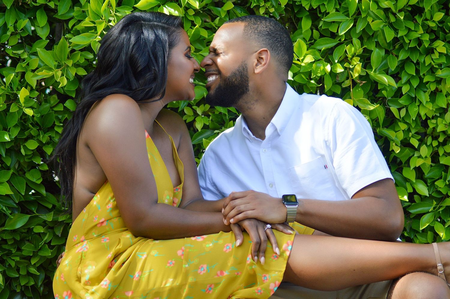 A couple shares a romantic moment, gazing into each other's eyes while holding hands outdoors near lush green foliage.