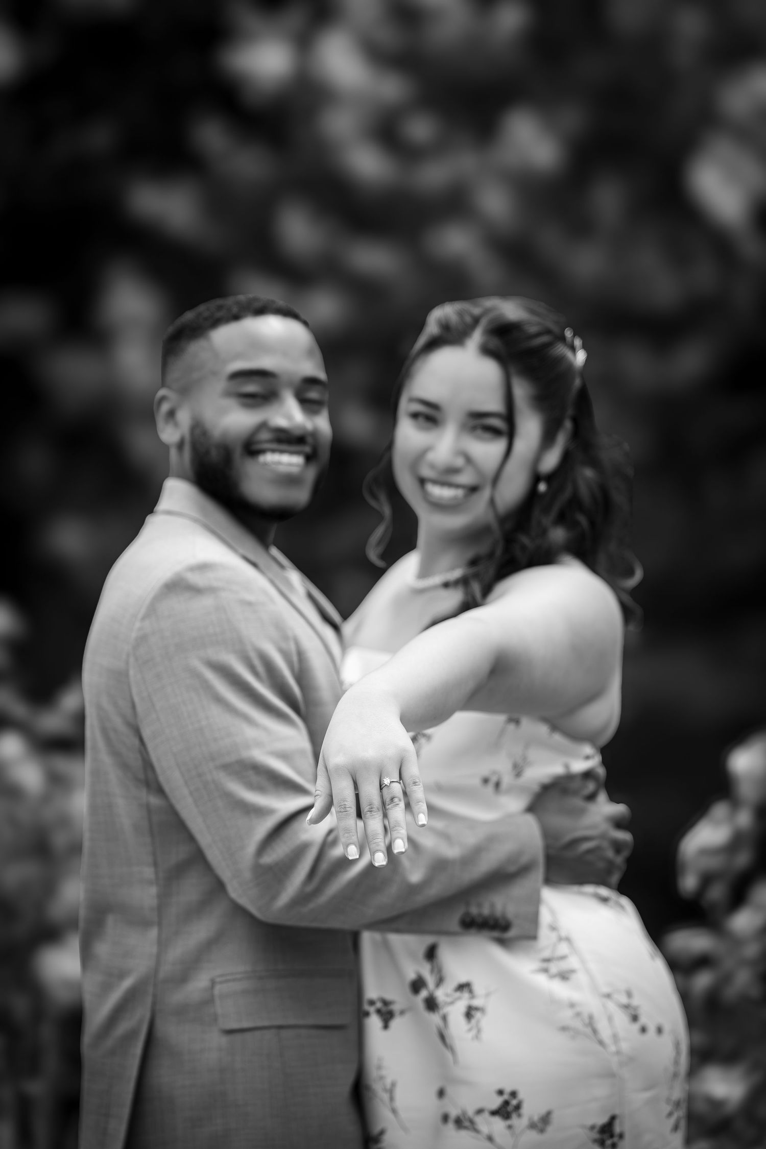 Black and white photo of a smiling couple embracing outdoors, woman displaying engagement ring, man in suit jacket.