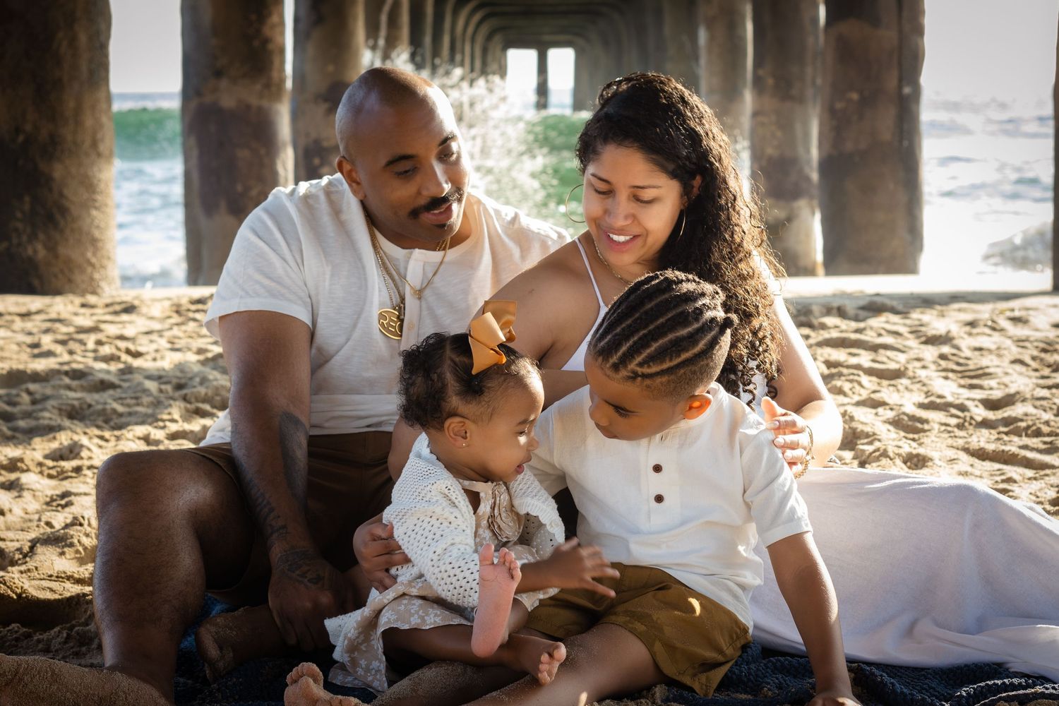 A family sits together on the beach under a pier during golden hour, sharing a tender moment in the warm sunlight.
